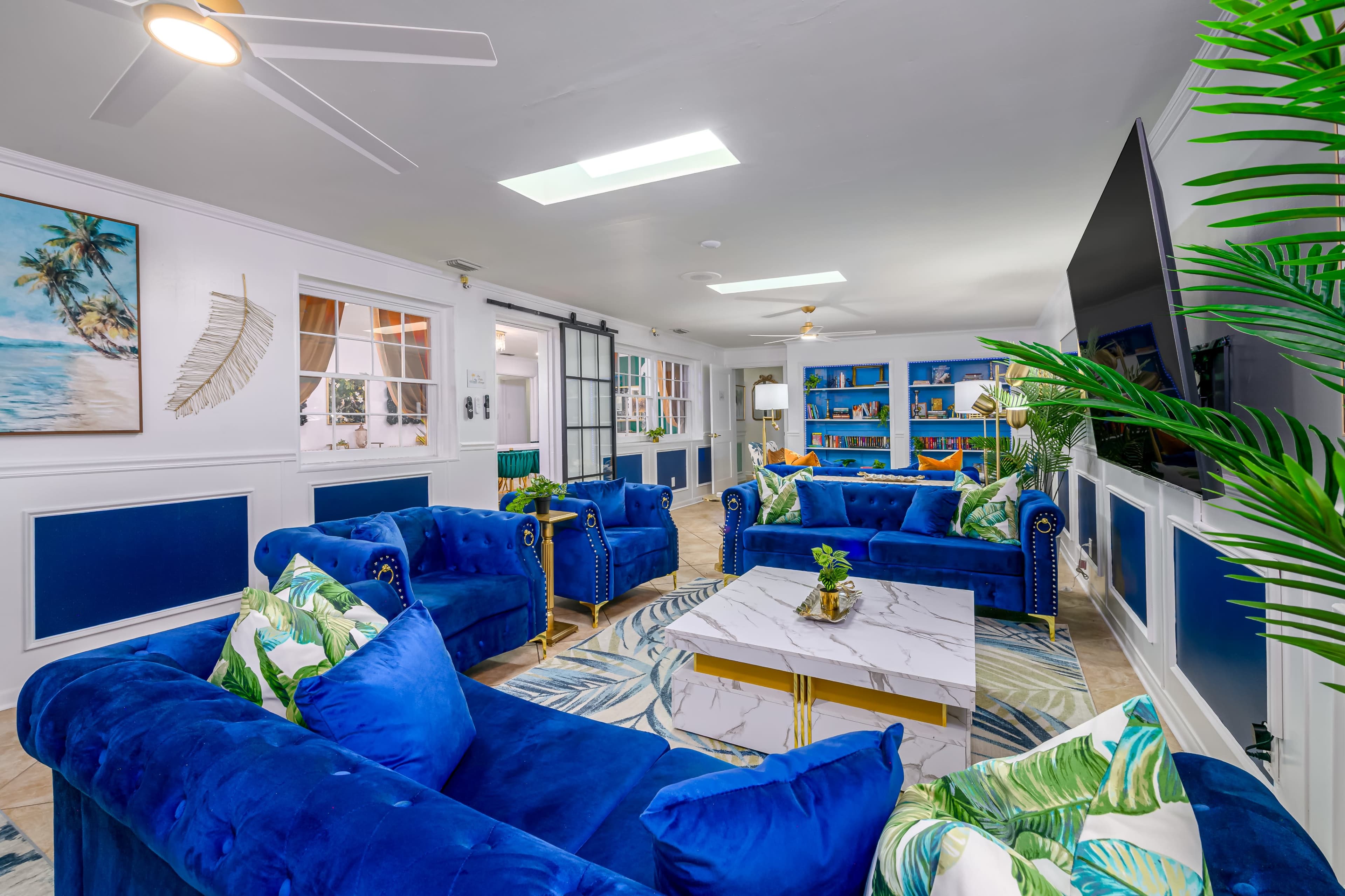 The image shows a modern living room with blue sofas, a marble coffee table, and tropical-themed decor.