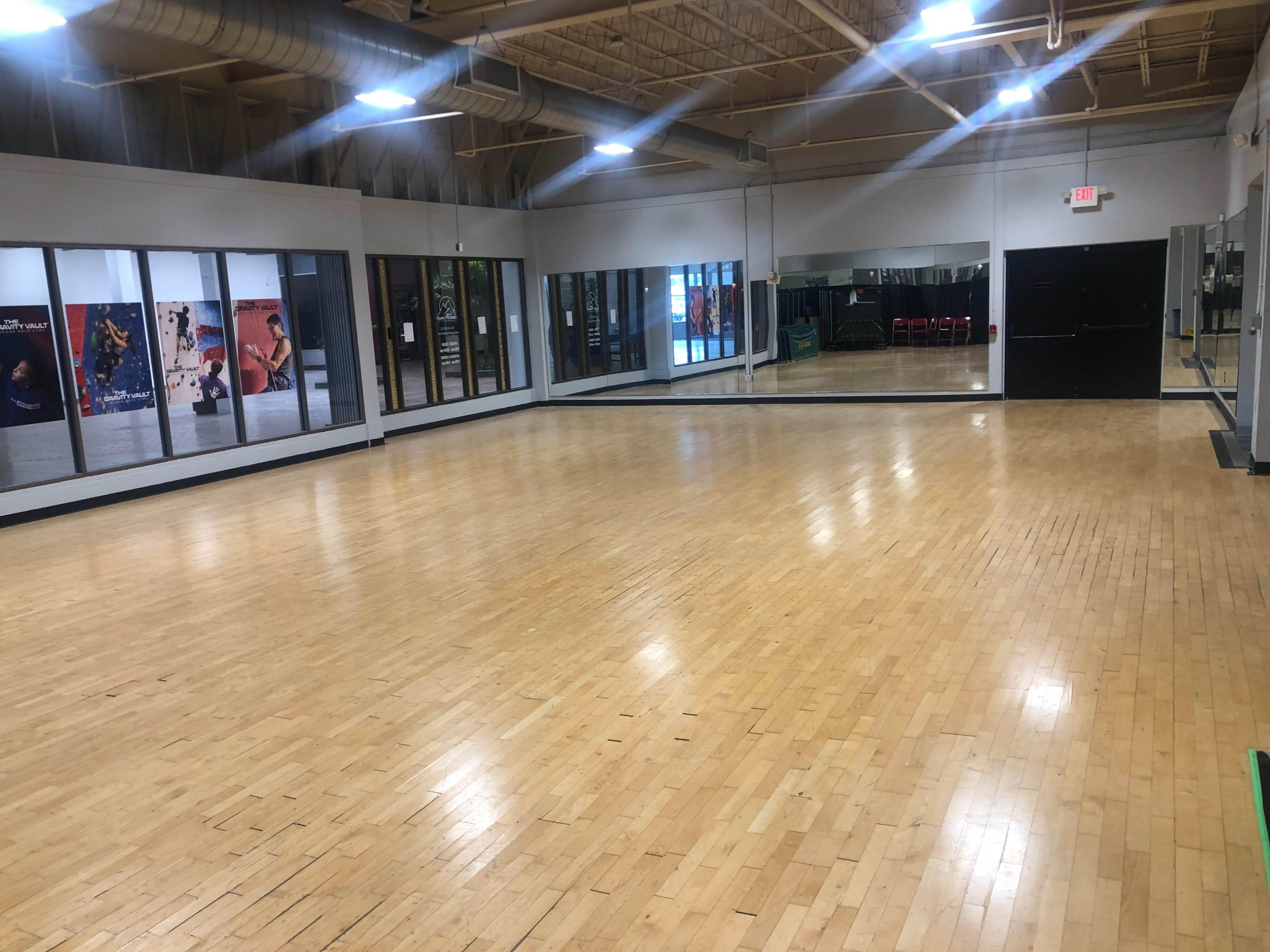 The image shows a spacious indoor studio with wooden flooring, large mirrors on one wall, and windows along another, reflecting a well-lit environment.