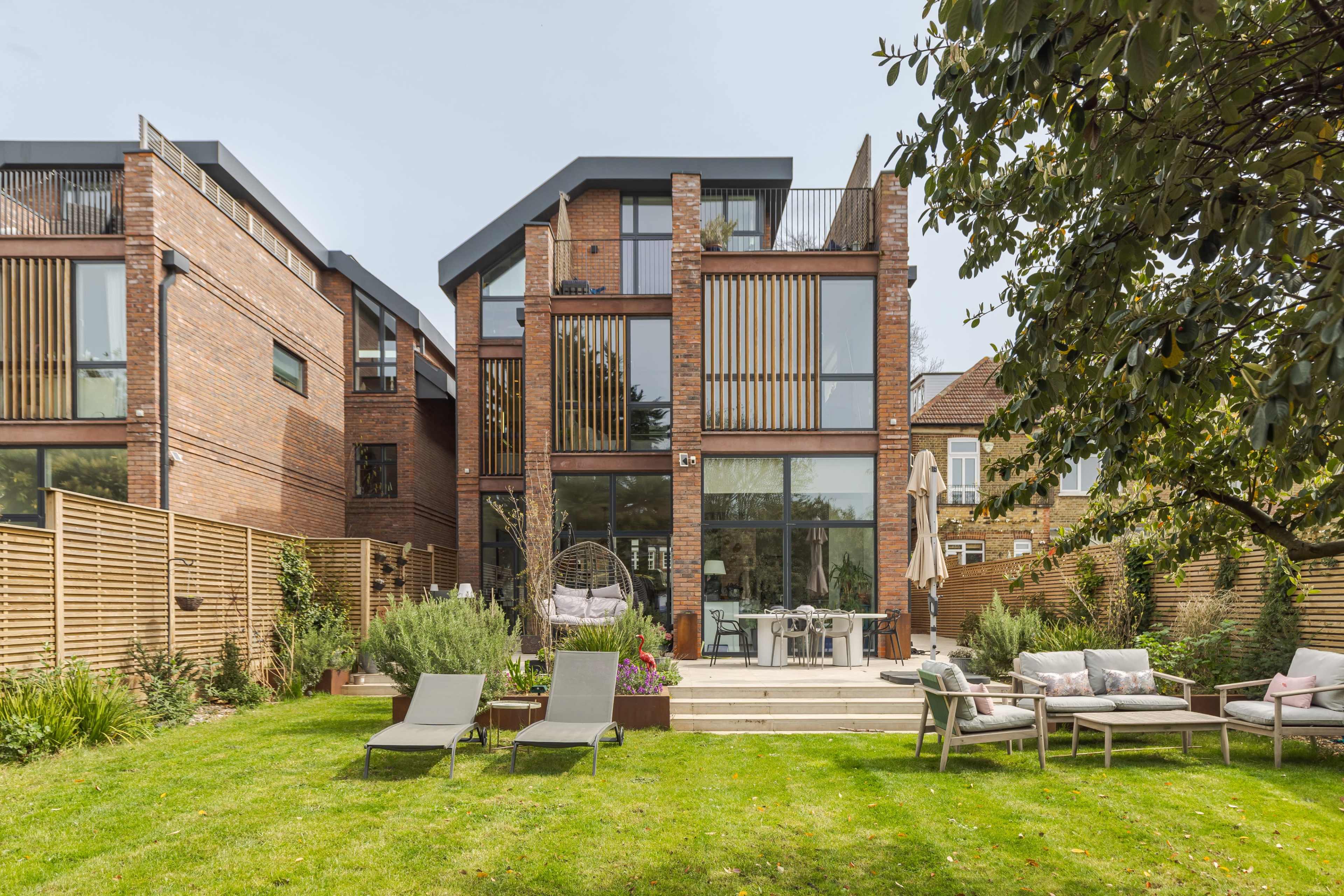 The image shows a modern brick house with large glass windows and a landscaped backyard featuring lounge chairs and outdoor furniture.