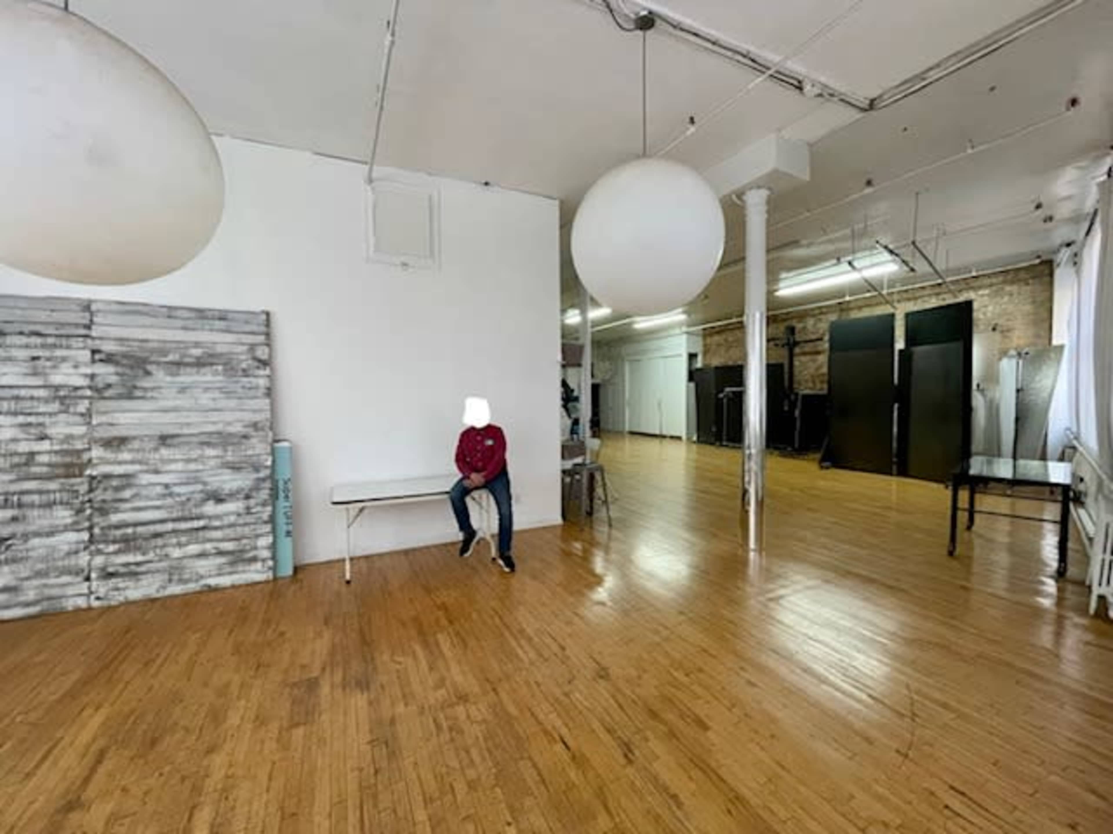 A person sits on a bench in a spacious, empty room with wooden floors and large white light fixtures.