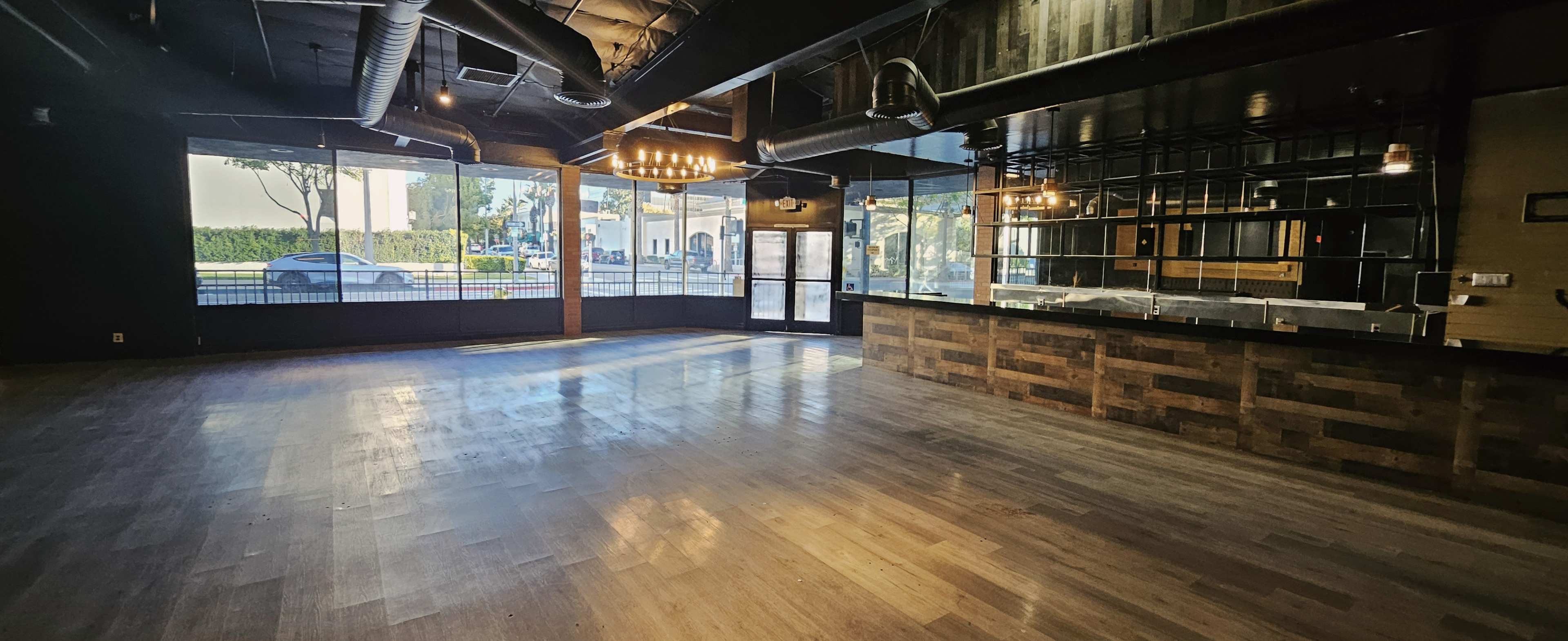 The image shows an empty interior space with wooden flooring, large windows, and a partially built bar area along one wall.