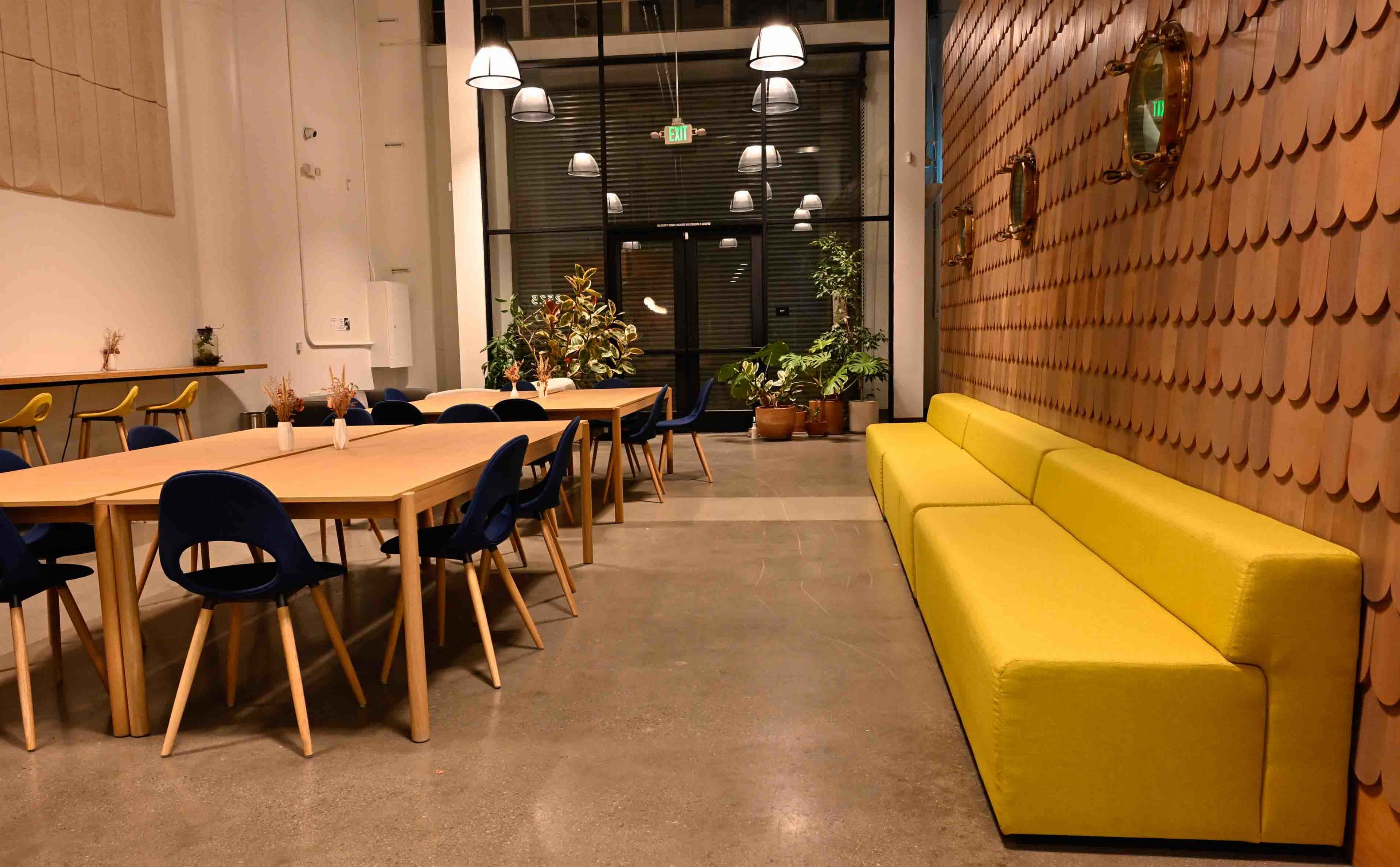 The image shows a modern interior space featuring a yellow sofa, a wooden dining table with chairs, and plants along the walls.