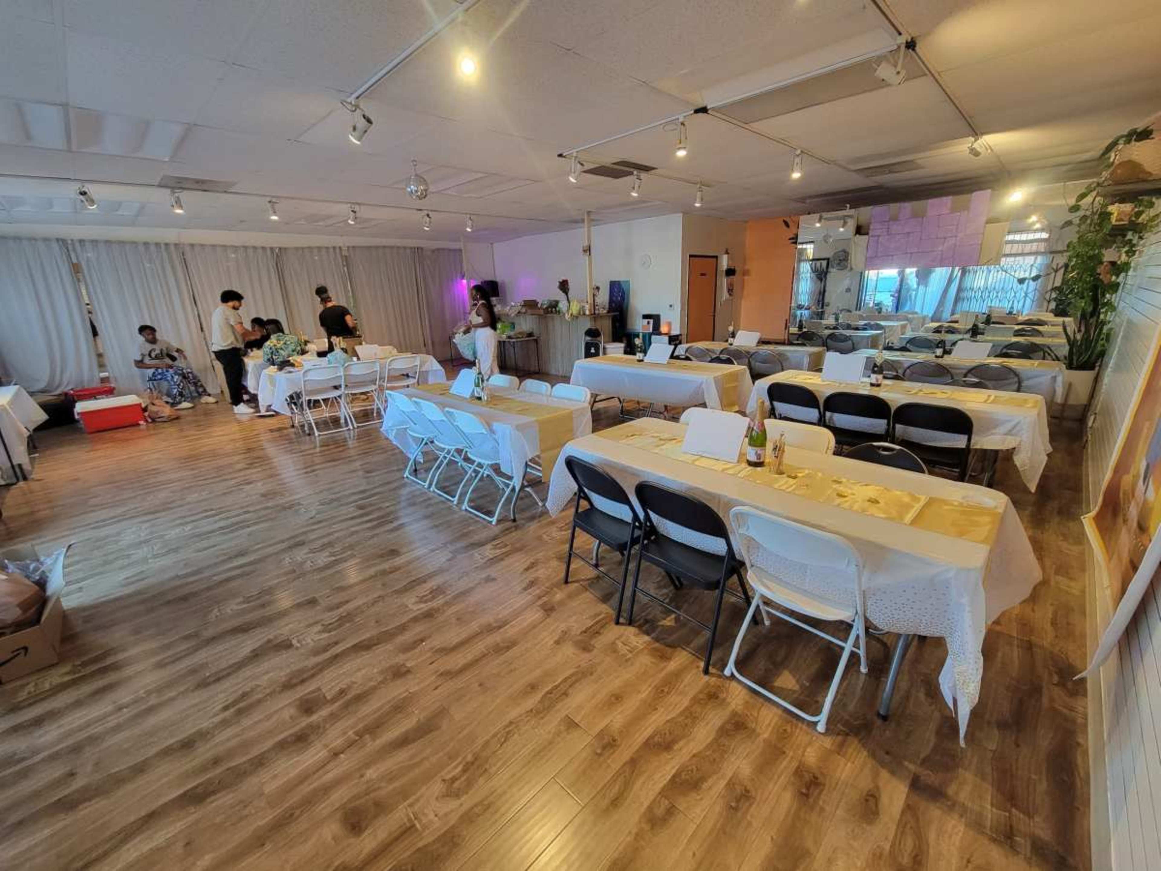 A spacious event room set up with several tables and chairs arranged for a gathering, with people engaged in various activities in the background.