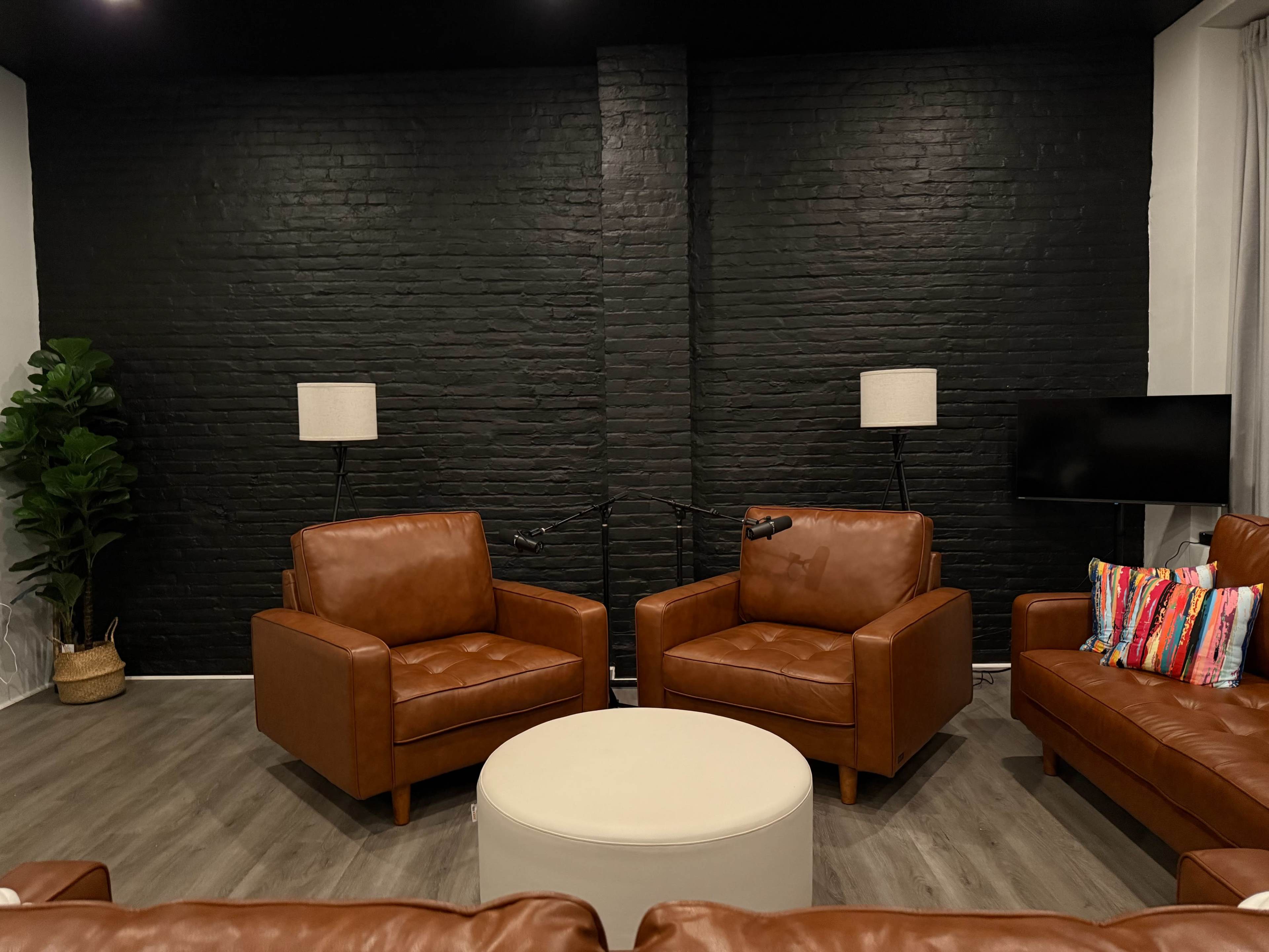 The image shows a modern living room with two brown leather armchairs, a round white coffee table, and a black brick wall in the background.