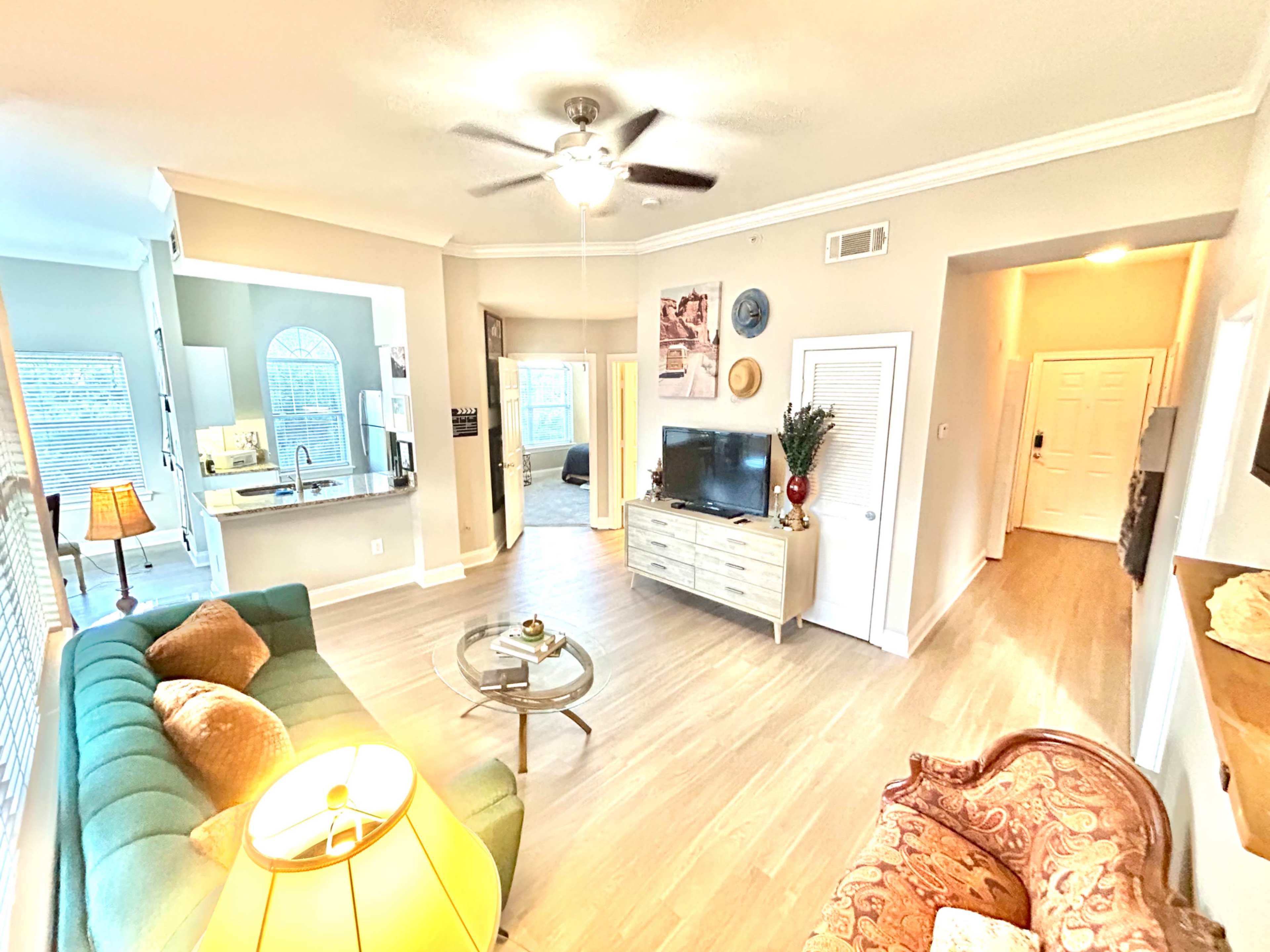 A spacious living room featuring a green sofa, a glass coffee table, and a ceiling fan, with an open layout leading to a kitchen and hallway.