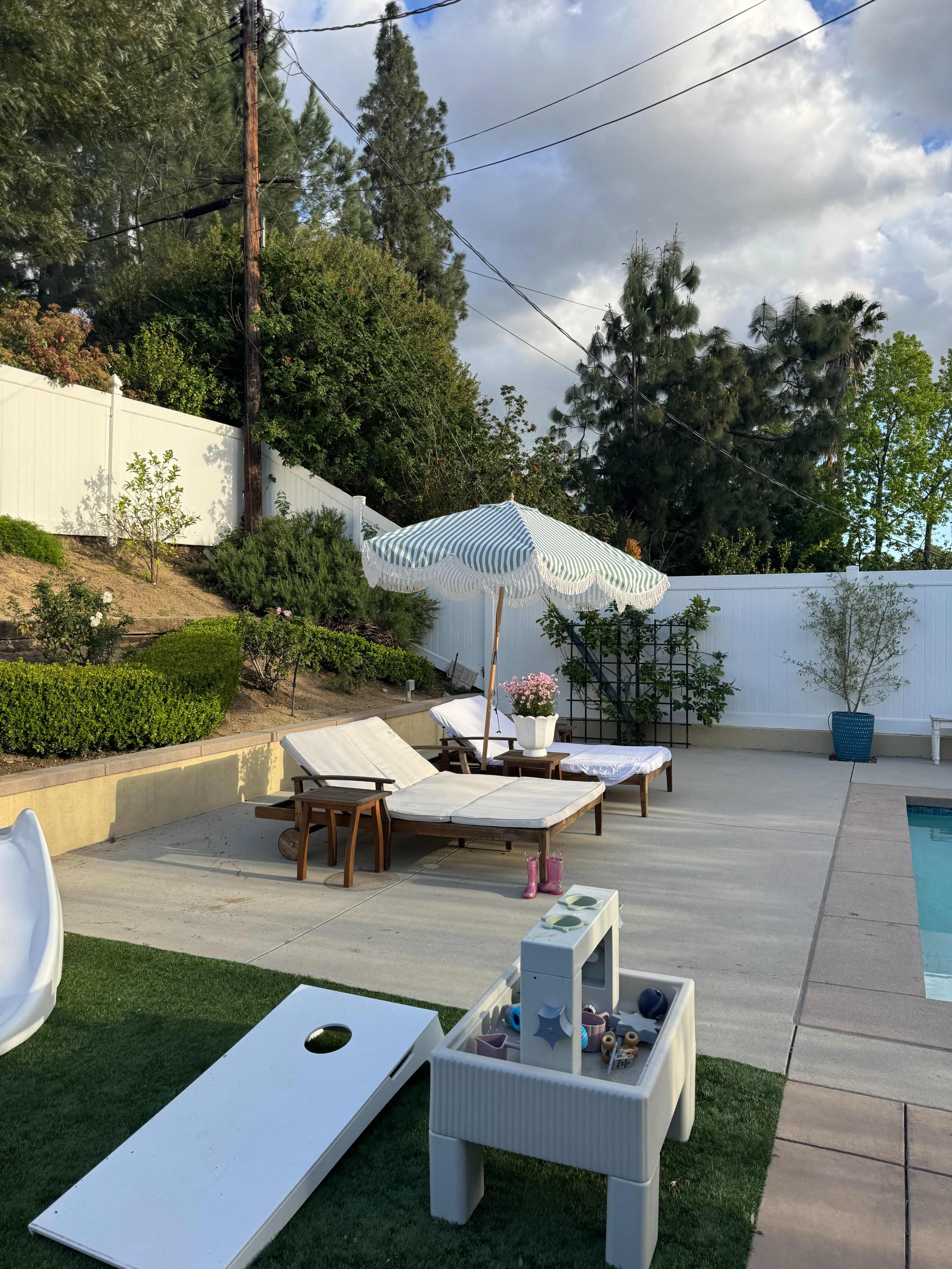 The image shows a pool area with two lounge chairs, a large umbrella, a cornhole game set up, and decorative plants against a white fence.