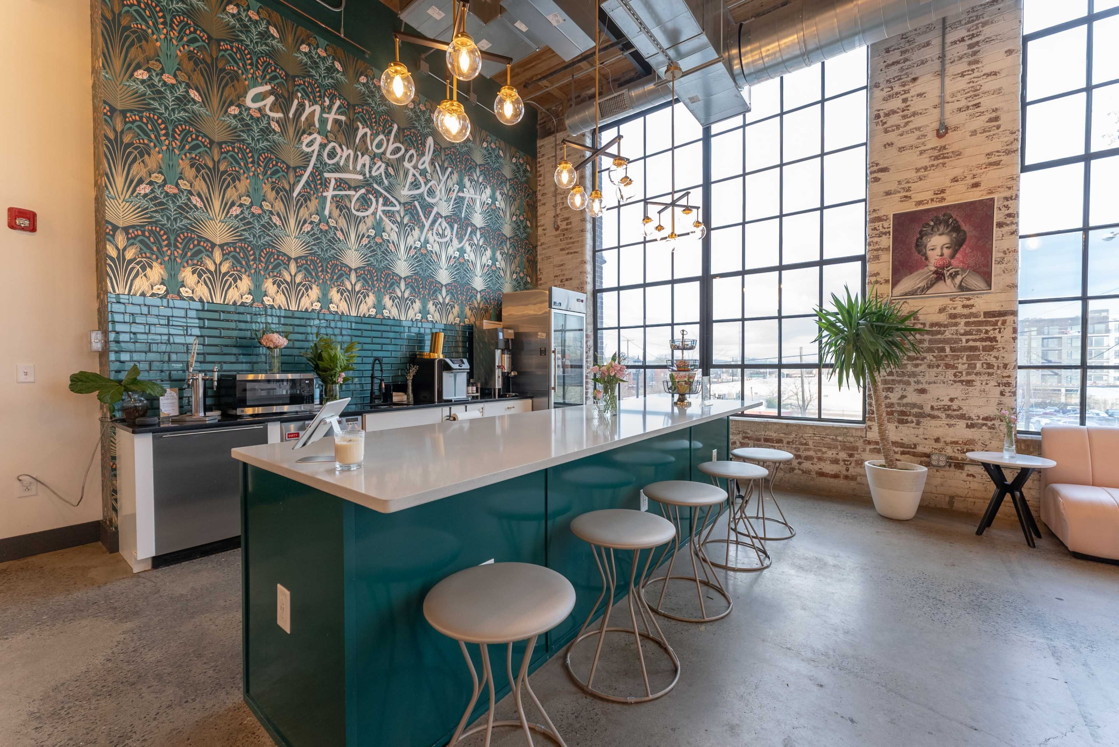 A modern kitchen with a large green island, white bar stools, and a decorative mural on the wall, illuminated by pendant lights and natural light from large windows.