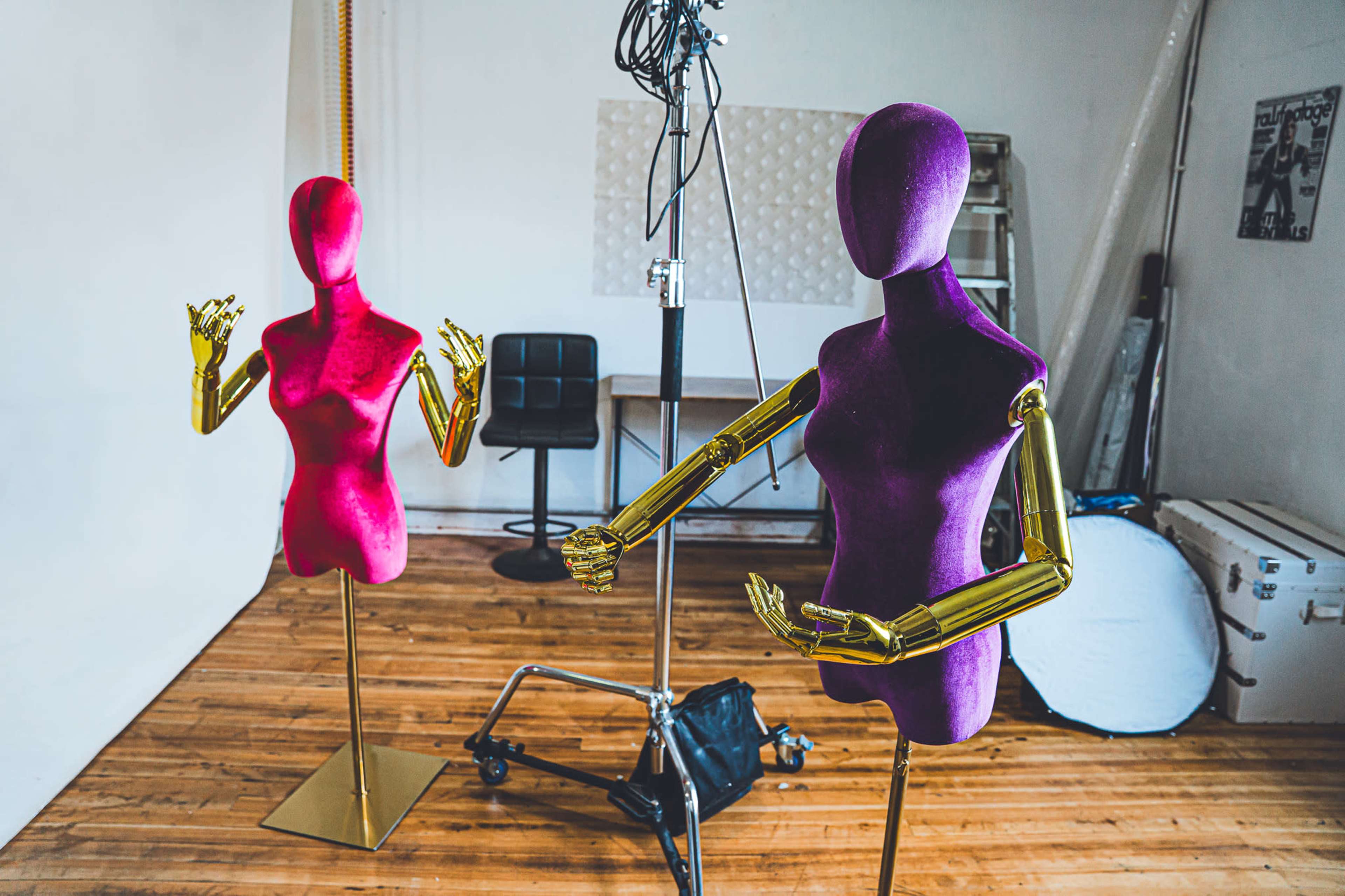 Two mannequins, one pink and one purple with golden hands, are positioned in a well-lit studio with a wooden floor and a backdrop.