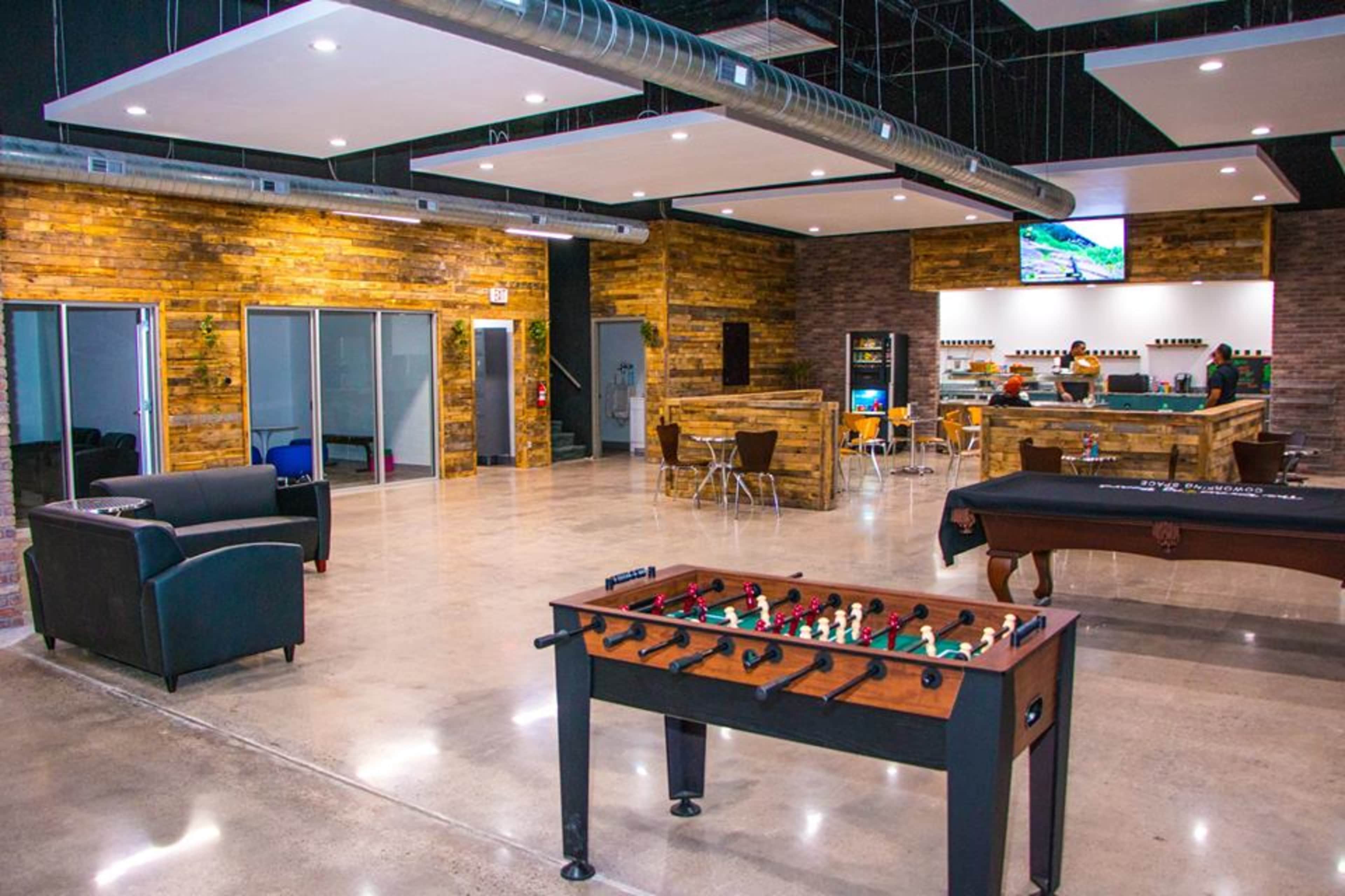 A spacious indoor recreational area featuring a foosball table, seating, a bar, and a screen displaying information.