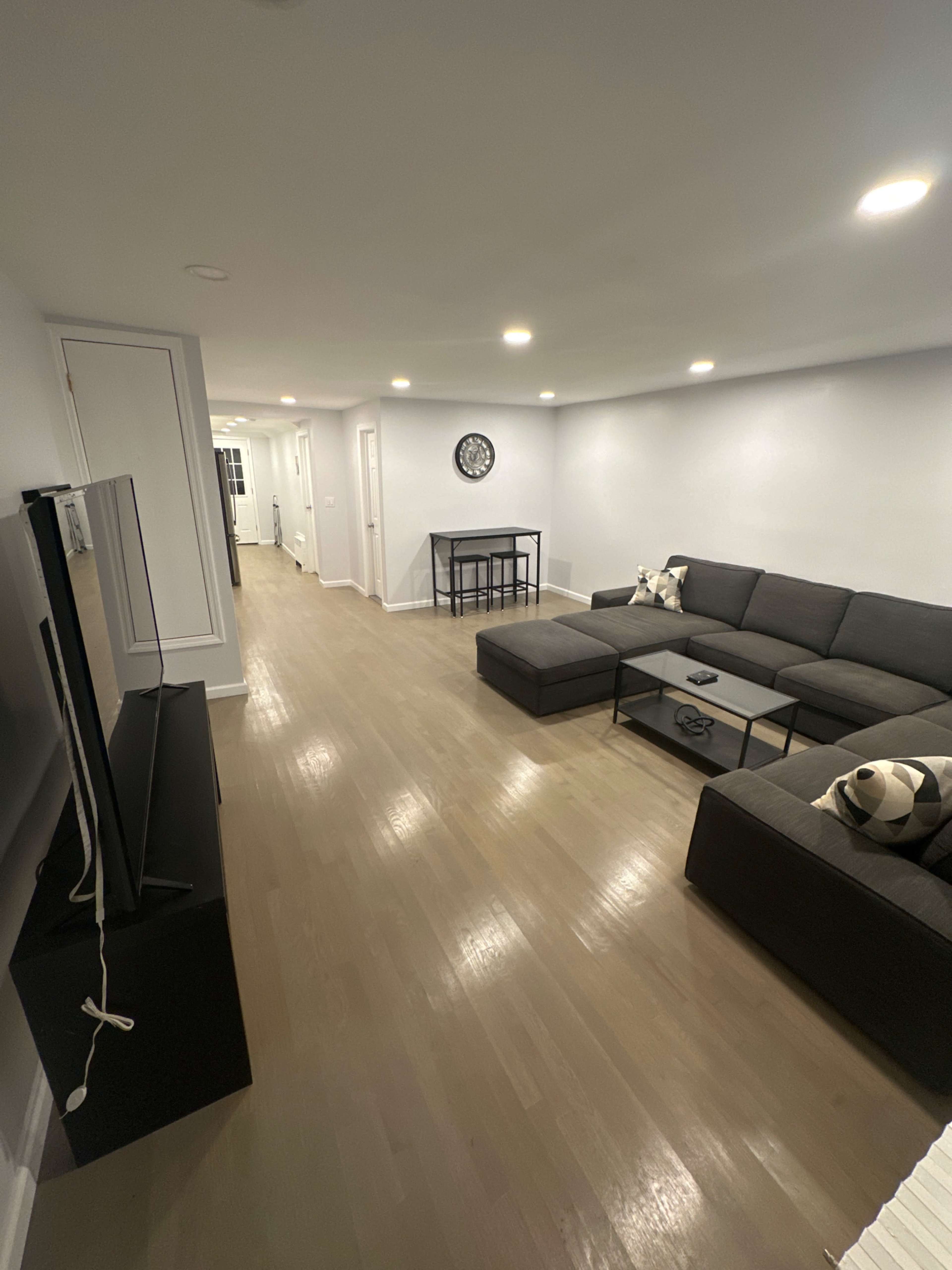 The image shows a modern, minimally furnished living room with a sectional sofa, a coffee table, and a small dining area in a well-lit space.