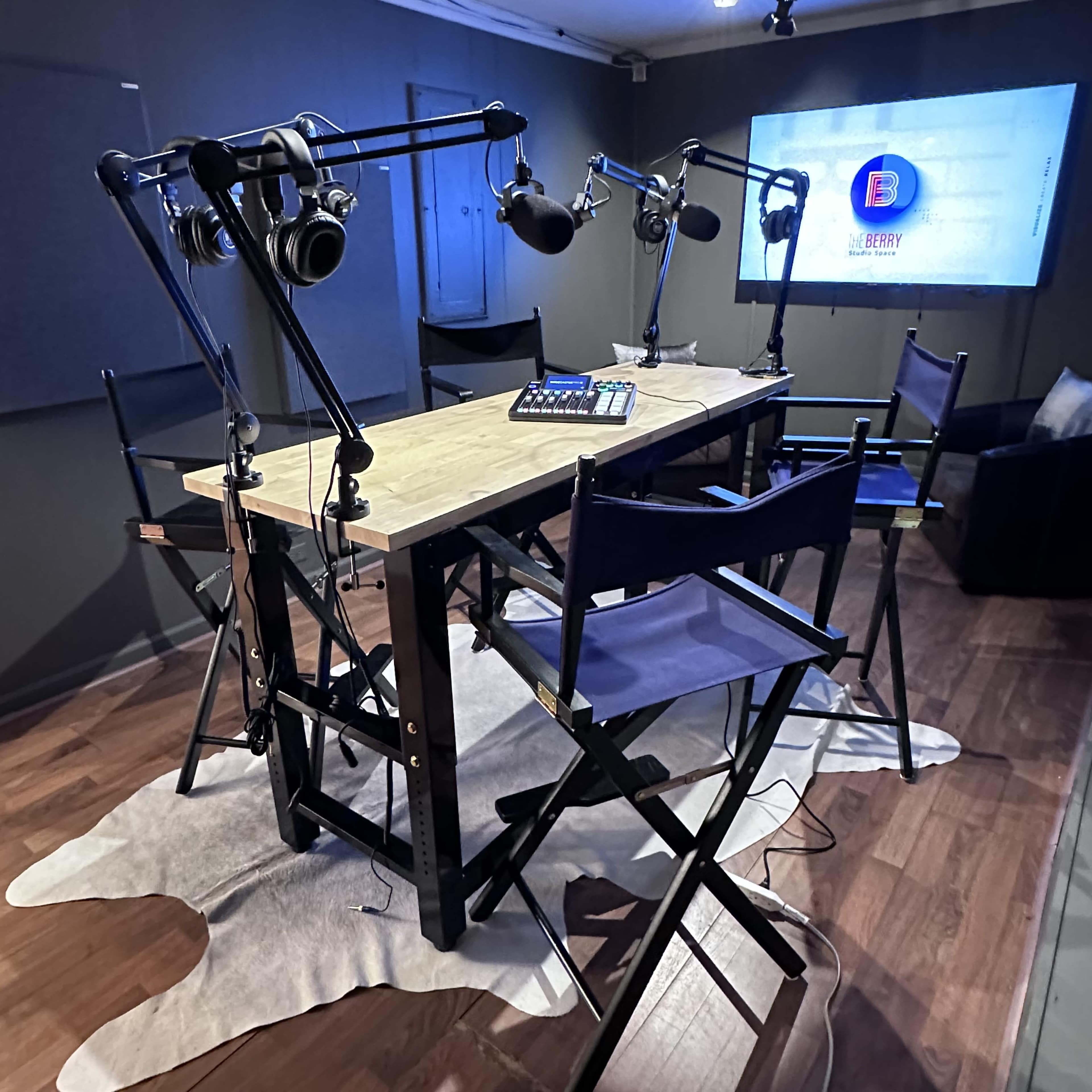The image shows a podcast recording setup featuring a wooden table with microphones and headphones, surrounded by several tall chairs on a cowhide rug.