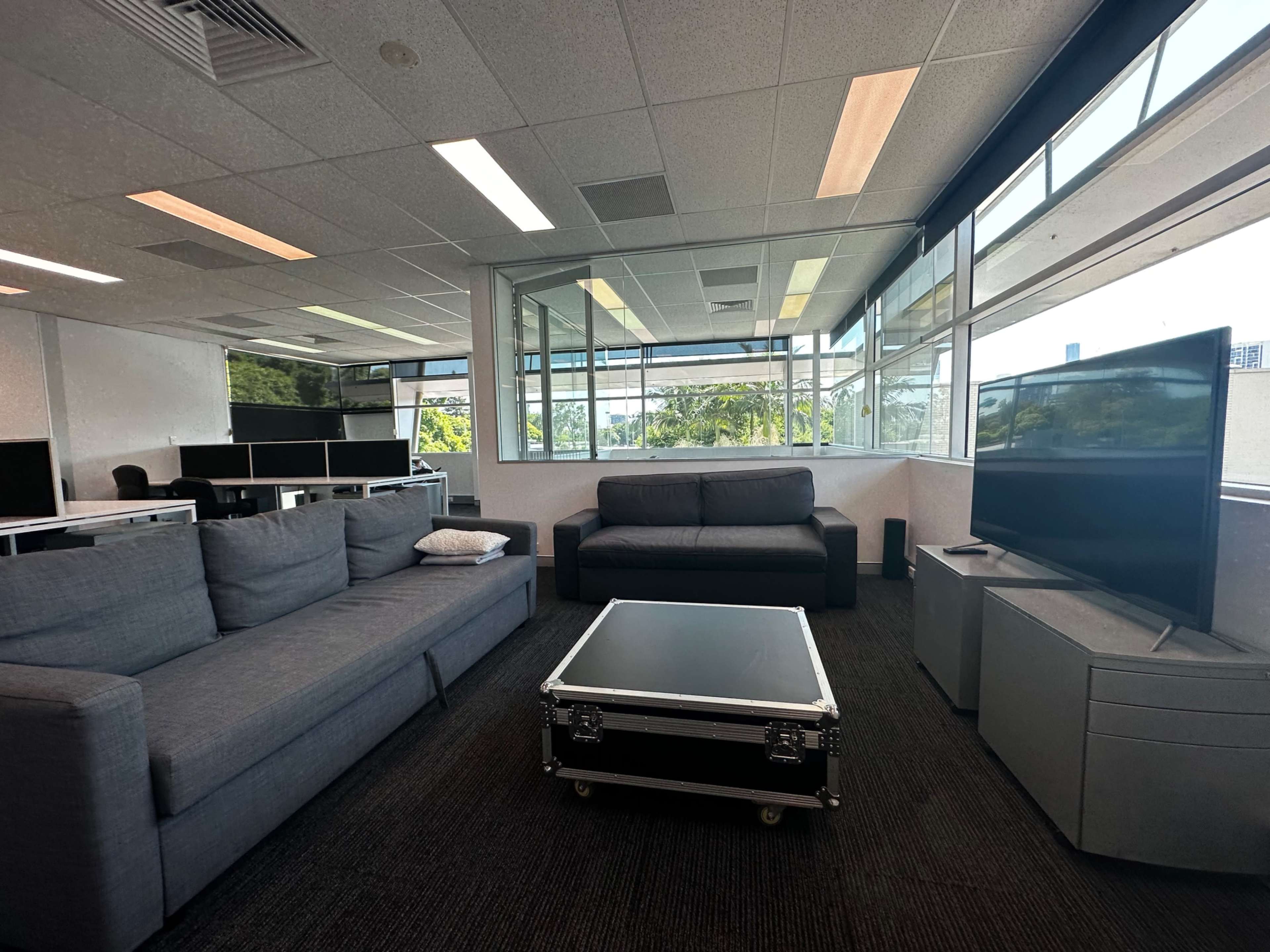 A modern office lounge area featuring a grey sectional couch, a coffee table, and a television, with large windows providing natural light and a view of the outdoors.