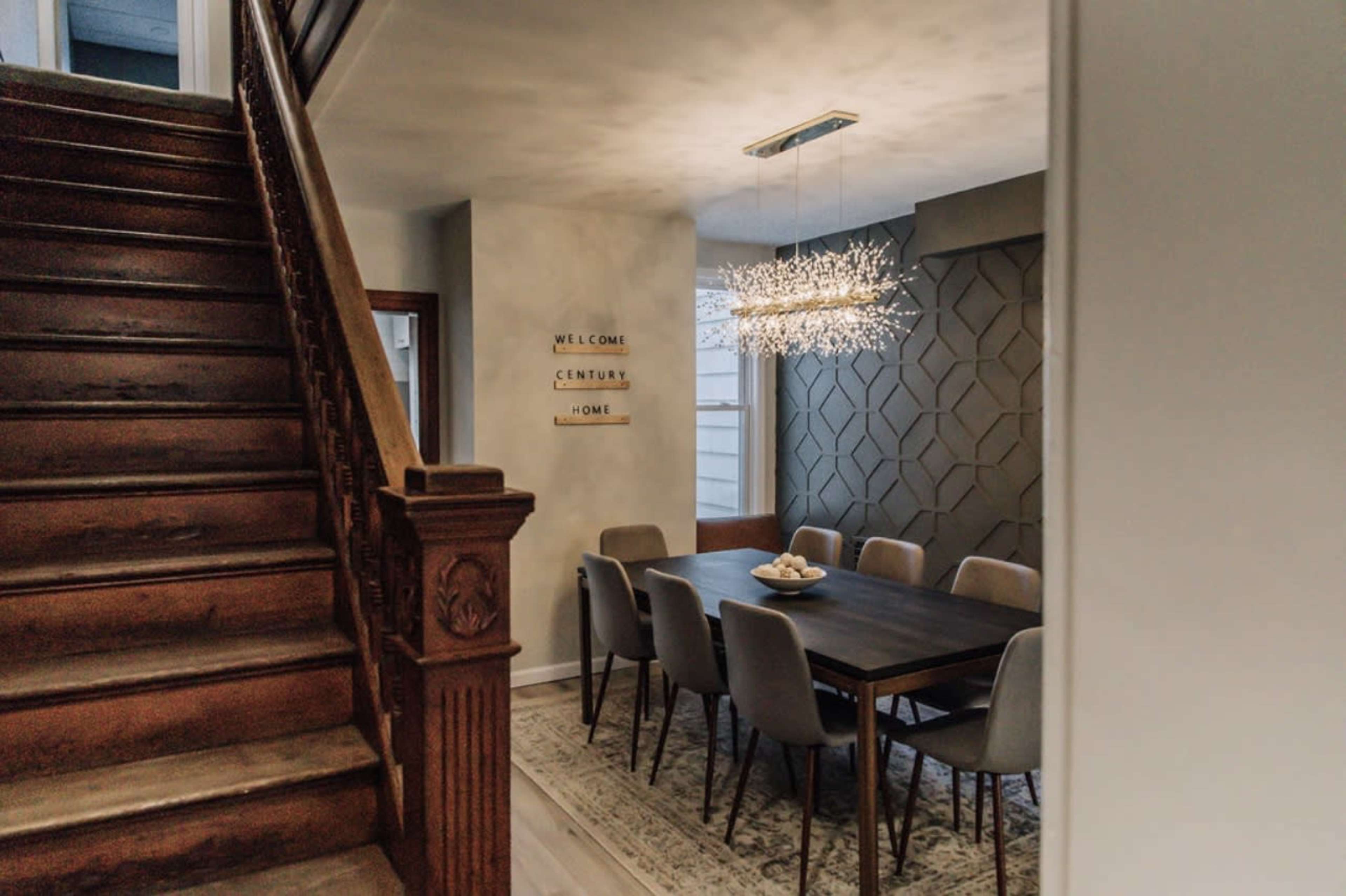 A wooden staircase leads to a dining area featuring a long table surrounded by gray chairs under a modern chandelier with an intricate design.