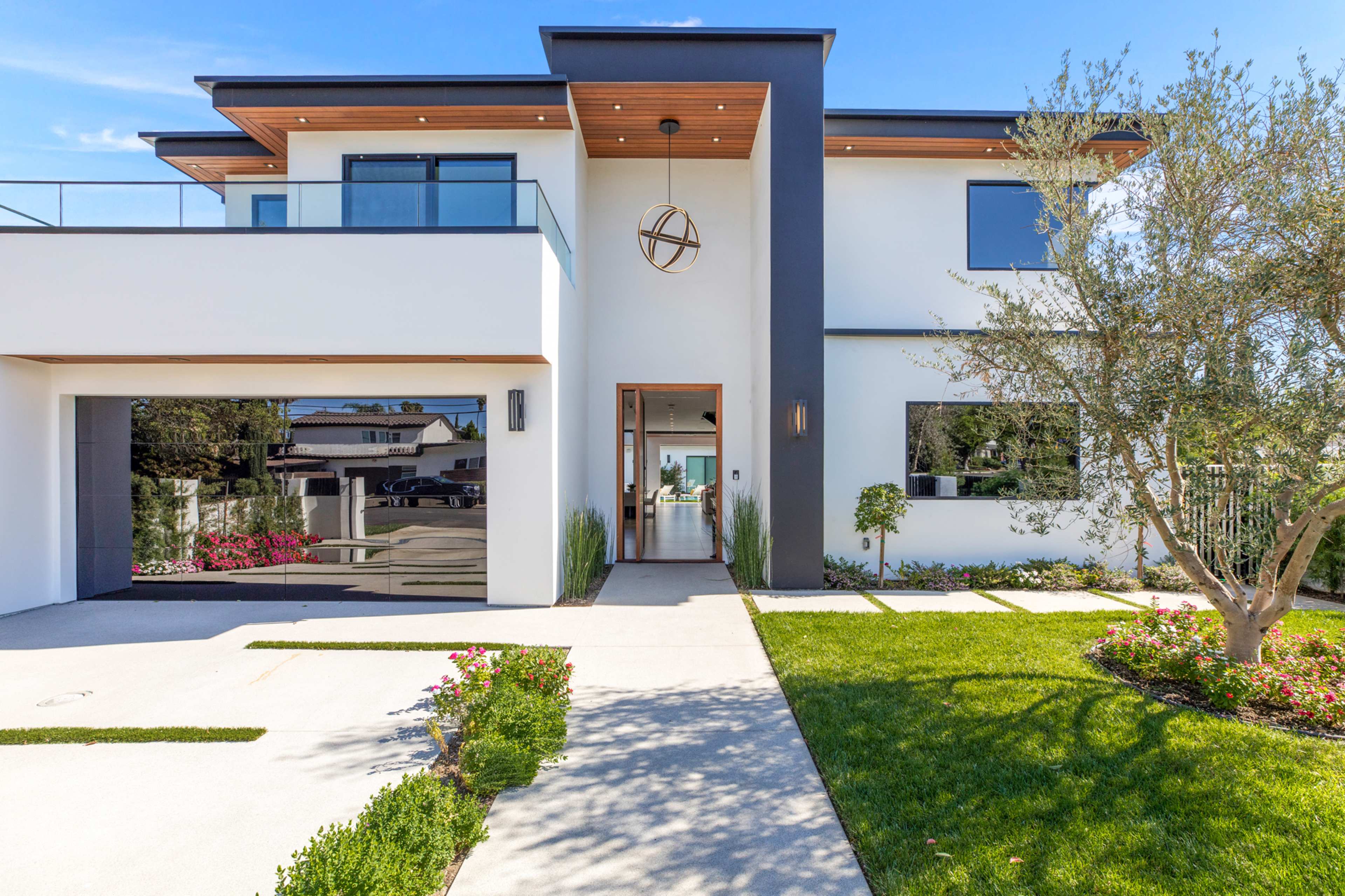 A modern two-story house with a striking facade, a concrete driveway, and landscaped gardens with flowers and greenery.