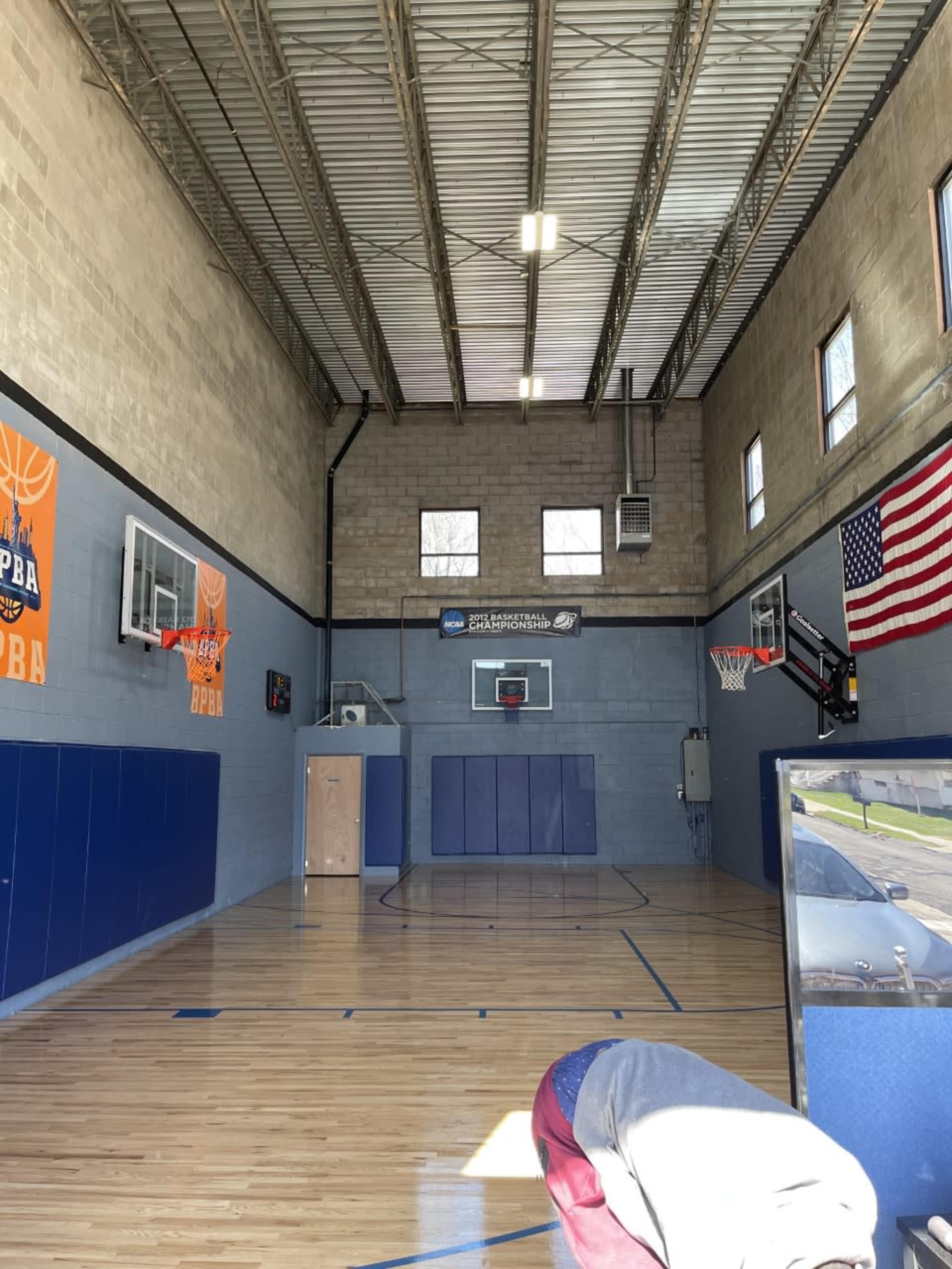 An indoor basketball court with two hoops, blue walls, wooden flooring, and an American flag displayed on one side.