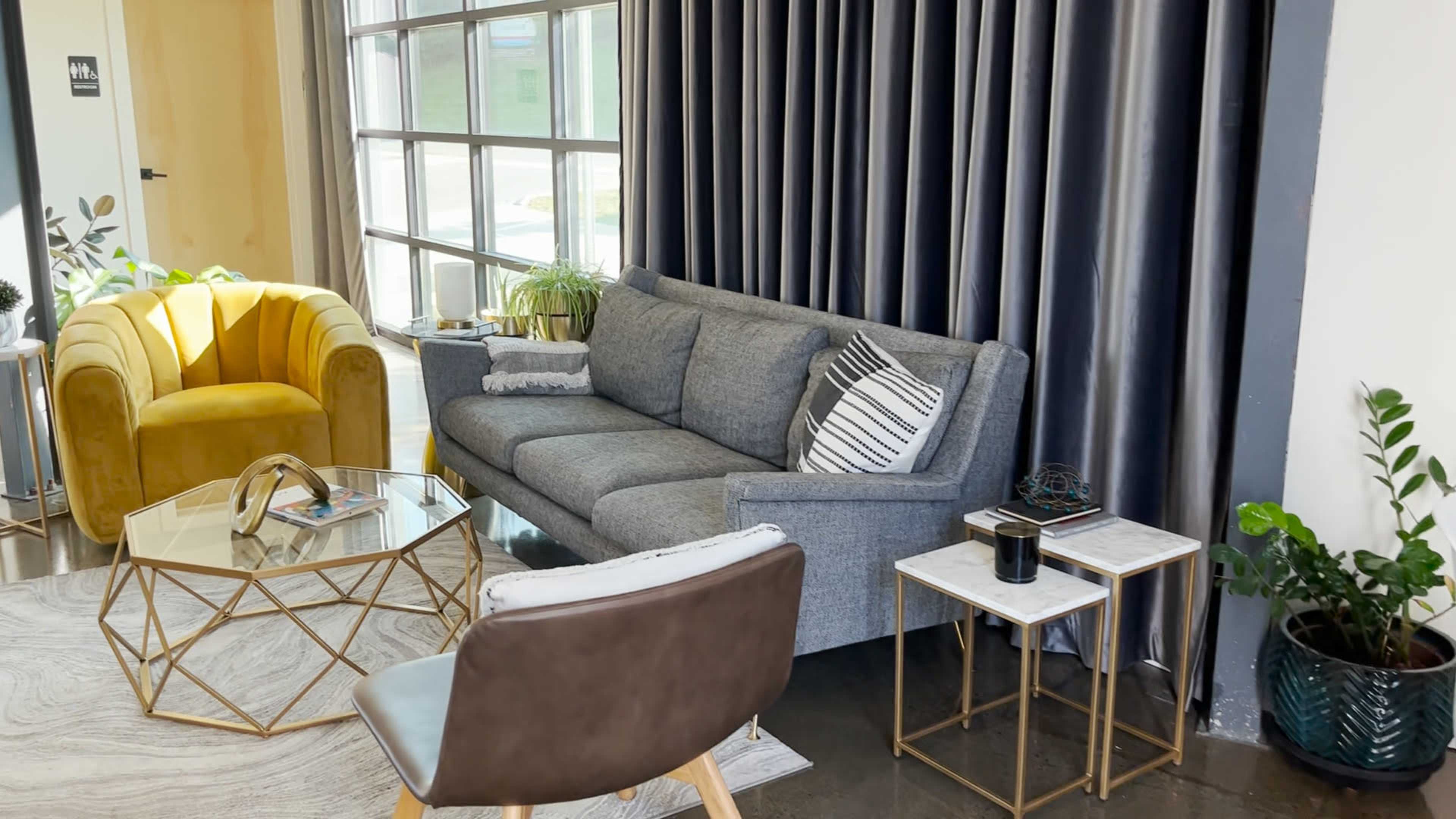 A modern living room with a gray sofa, a yellow armchair, a glass coffee table, and various plants in decorative arrangements.