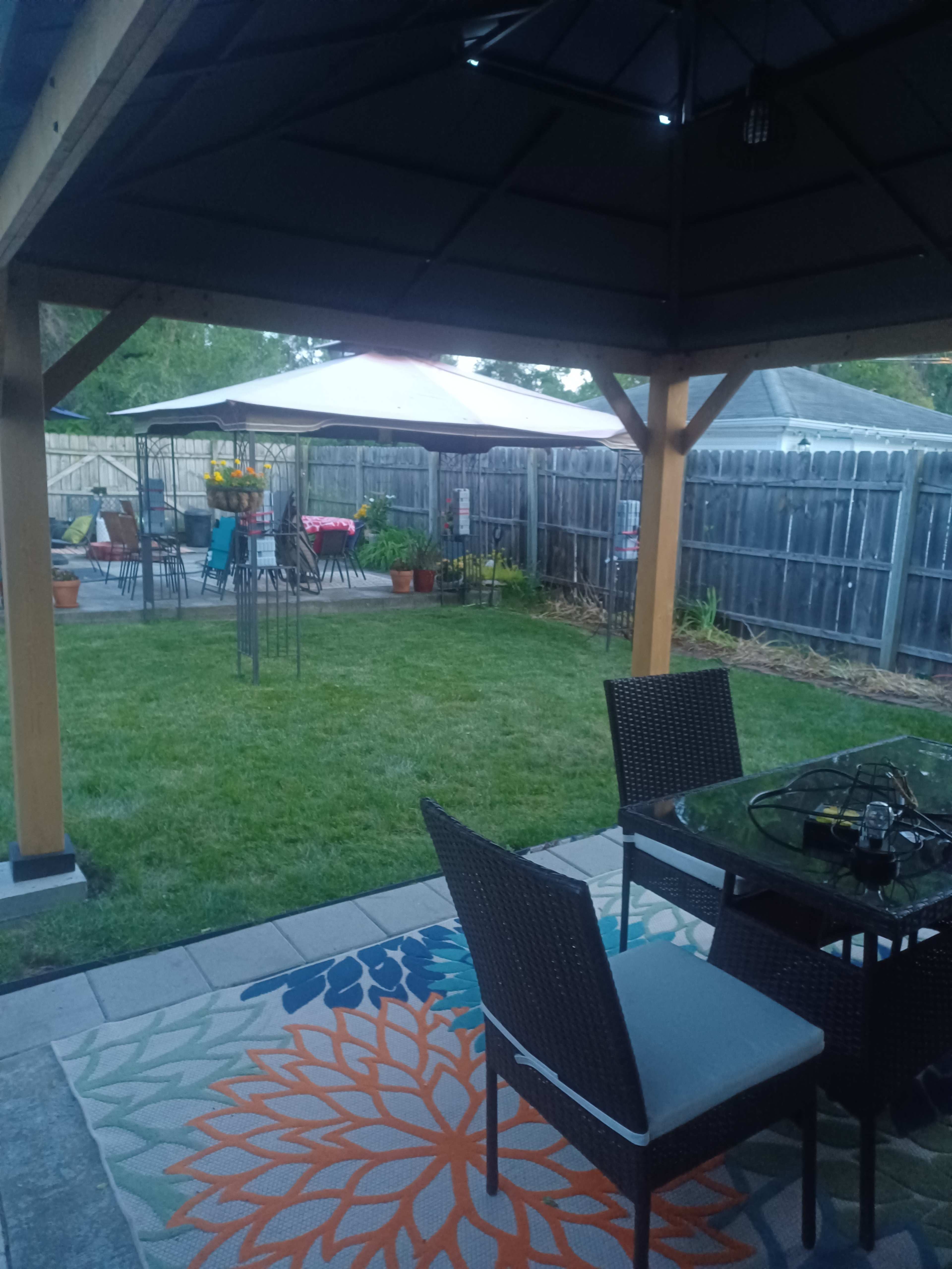 A backyard seating area features a table and chairs under a gazebo, overlooking a grassy lawn with additional seating and potted plants in the background.