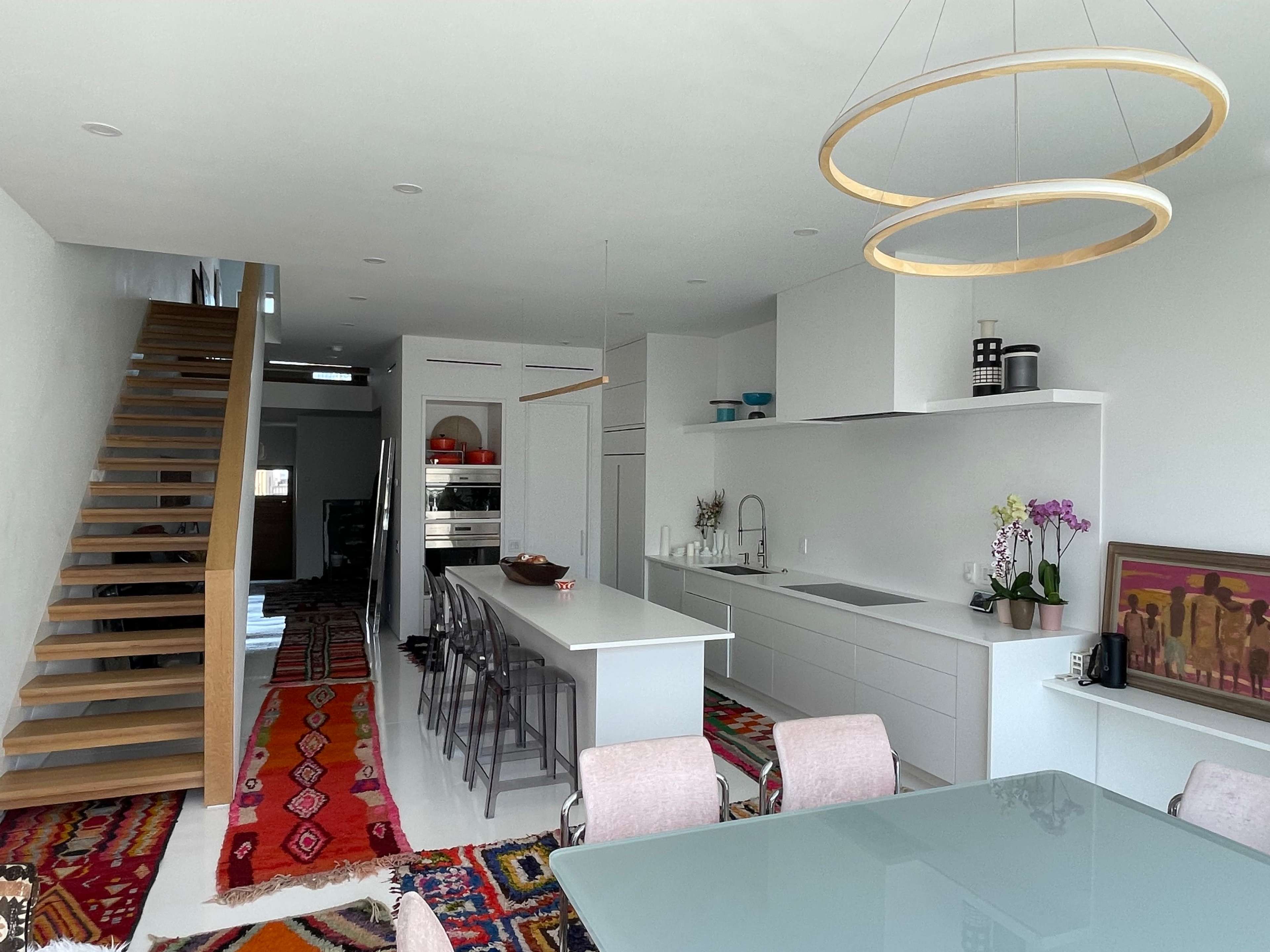 A modern open-concept kitchen and dining area with a staircase in the background and colorful rugs on the floor.
