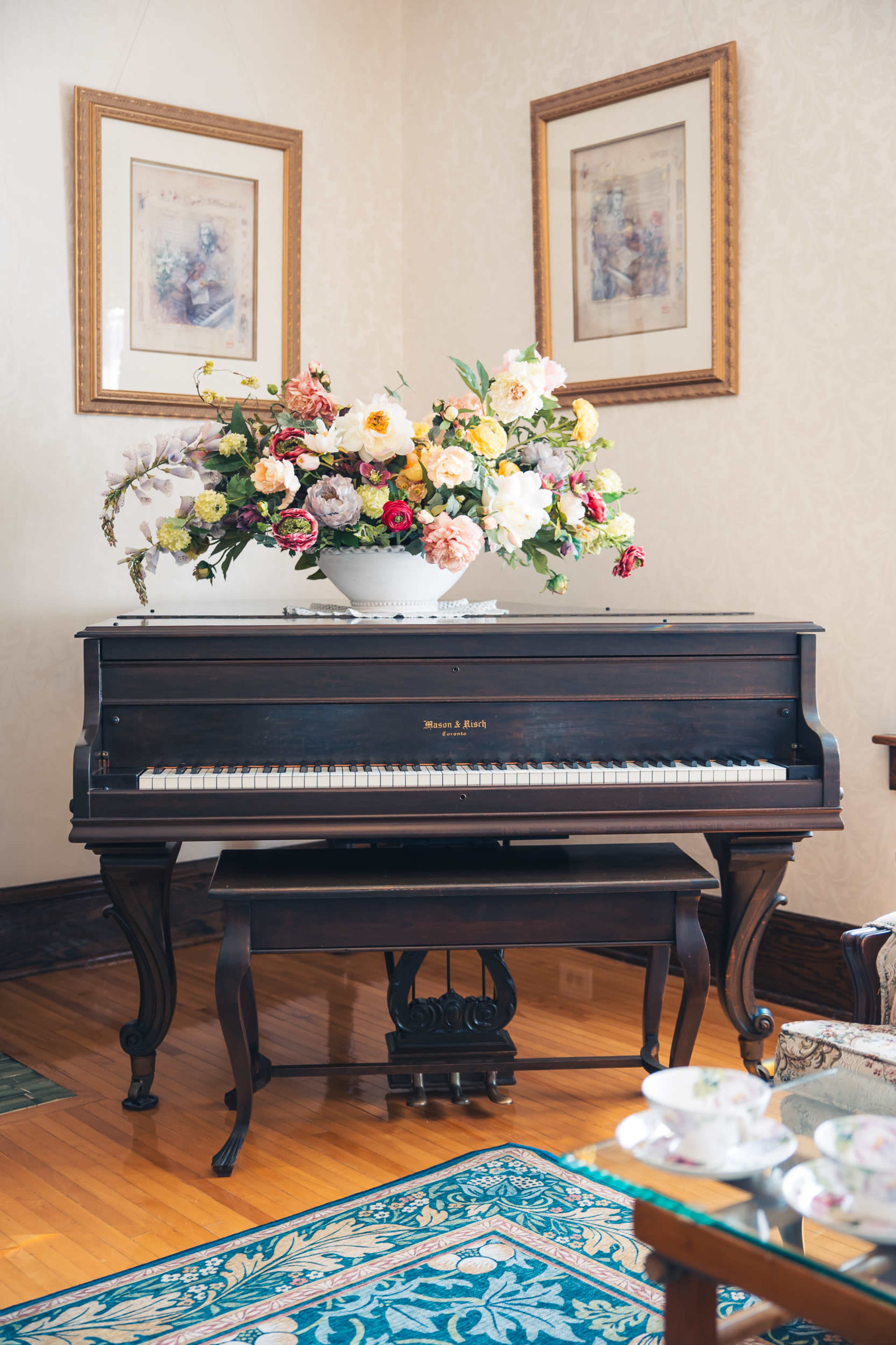 A grand piano with a floral arrangement on top sits in a well-decorated room with light-colored walls and wooden flooring.