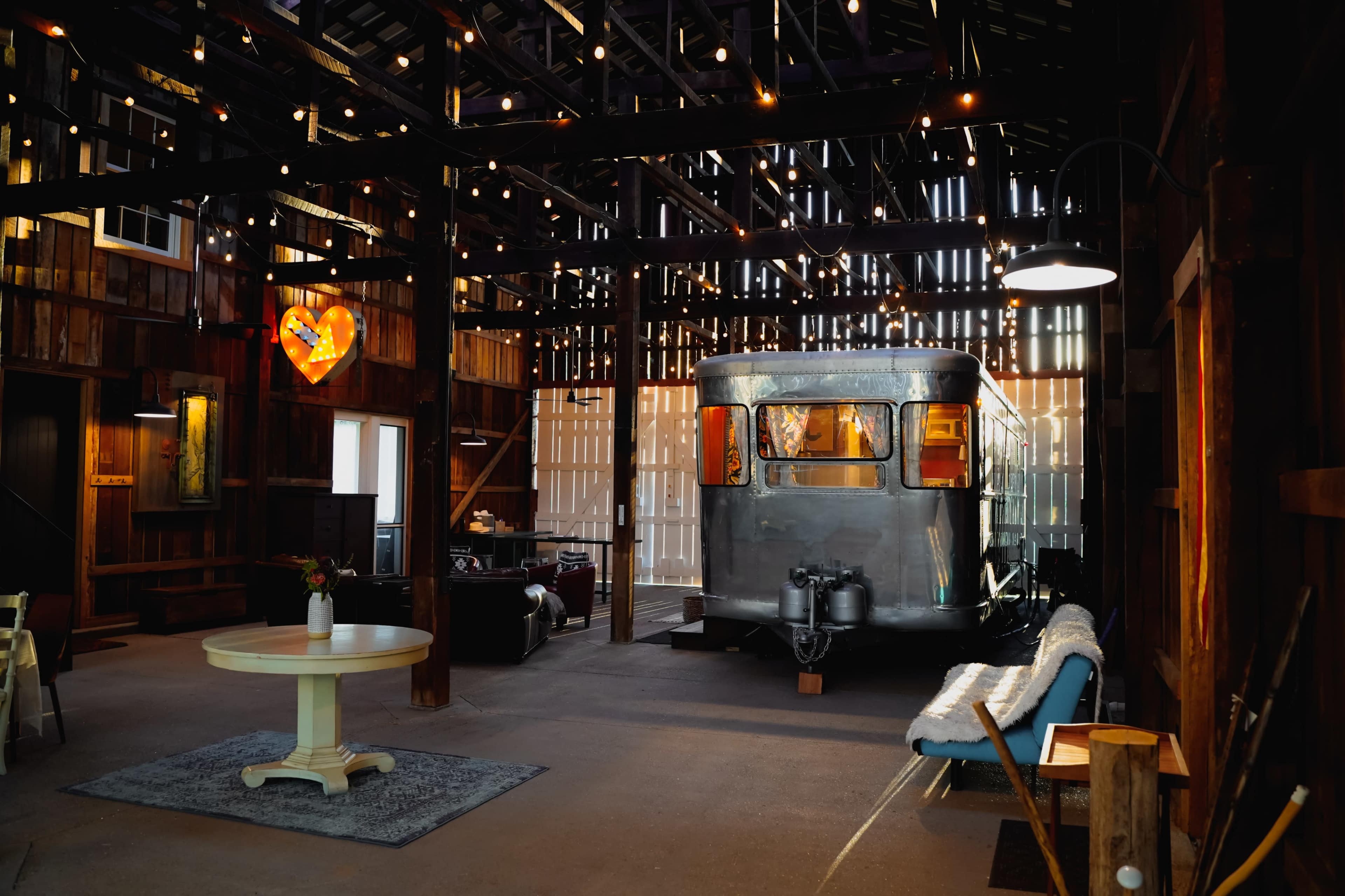 The interior of a rustic barn features an vintage silver trailer positioned prominently among wooden beams and warm lighting.