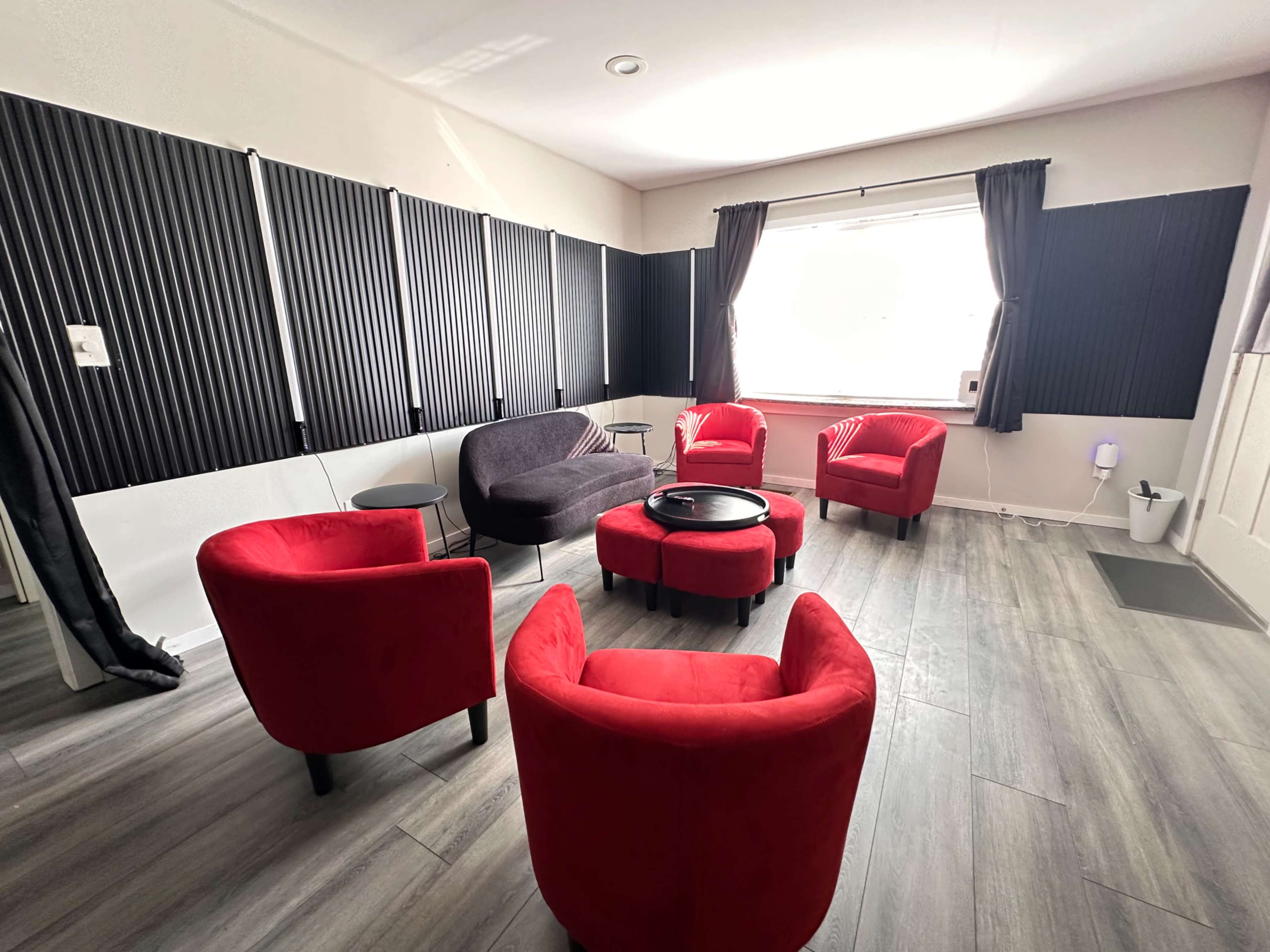 The image shows a minimalist room featuring five red chairs arranged around a black circular table, with gray walls and acoustic panels lining the windows.