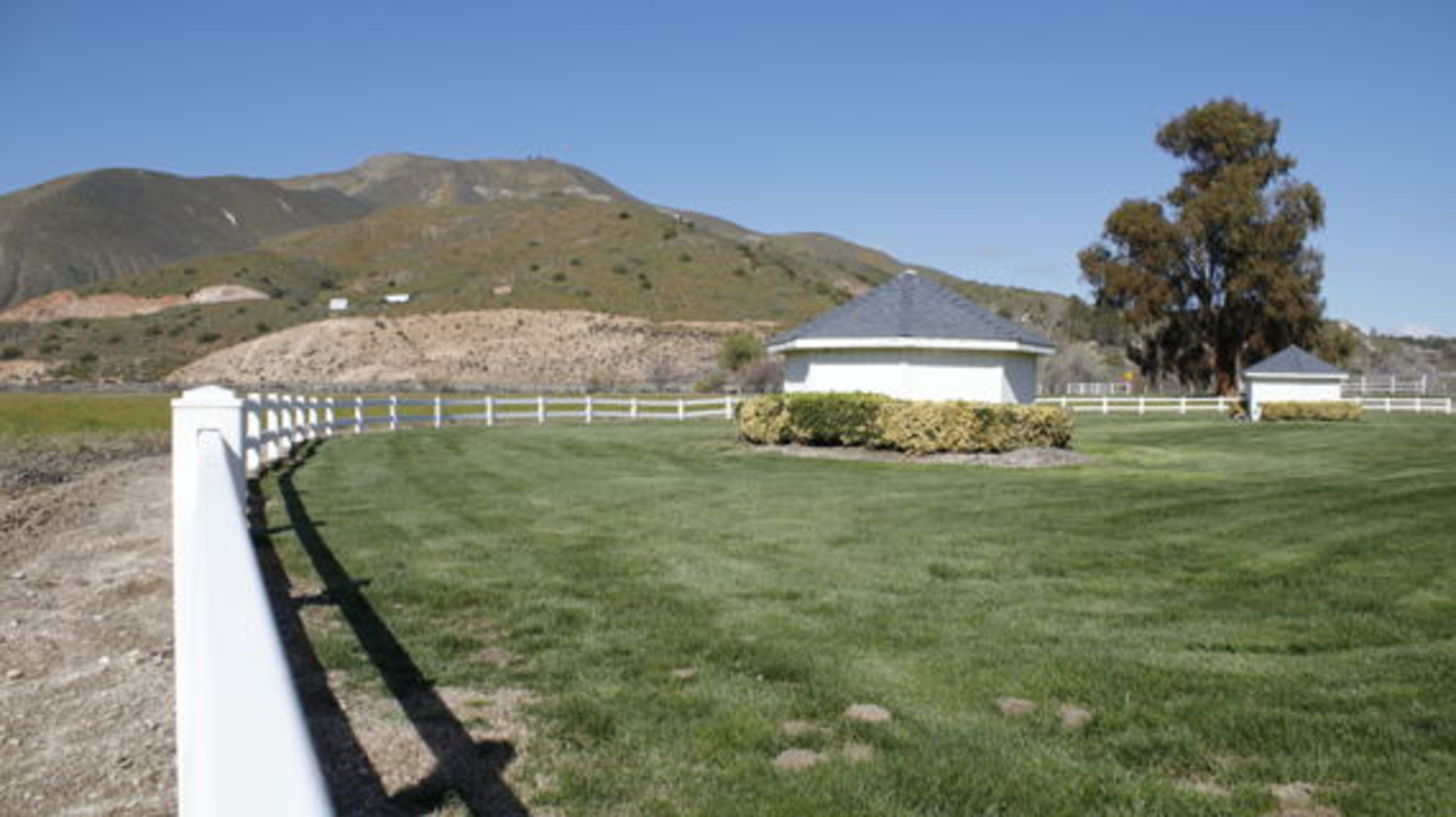 A white fence surrounds a well-maintained grassy area featuring two small buildings near a hill in the background.