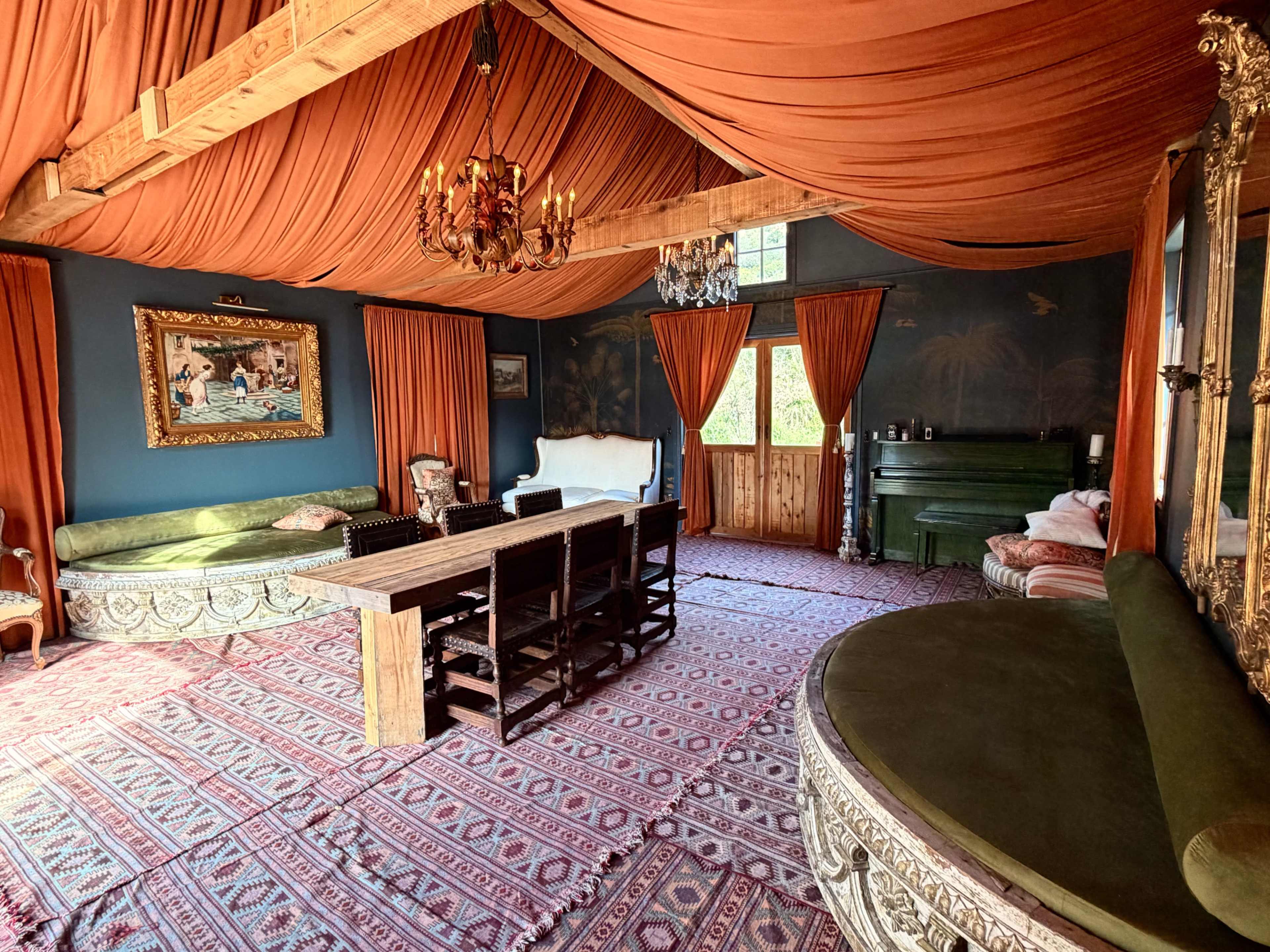The image shows a room with orange draped fabric covering the ceiling, a long wooden dining table surrounded by black chairs, and a green piano against a wall adorned with a painting.