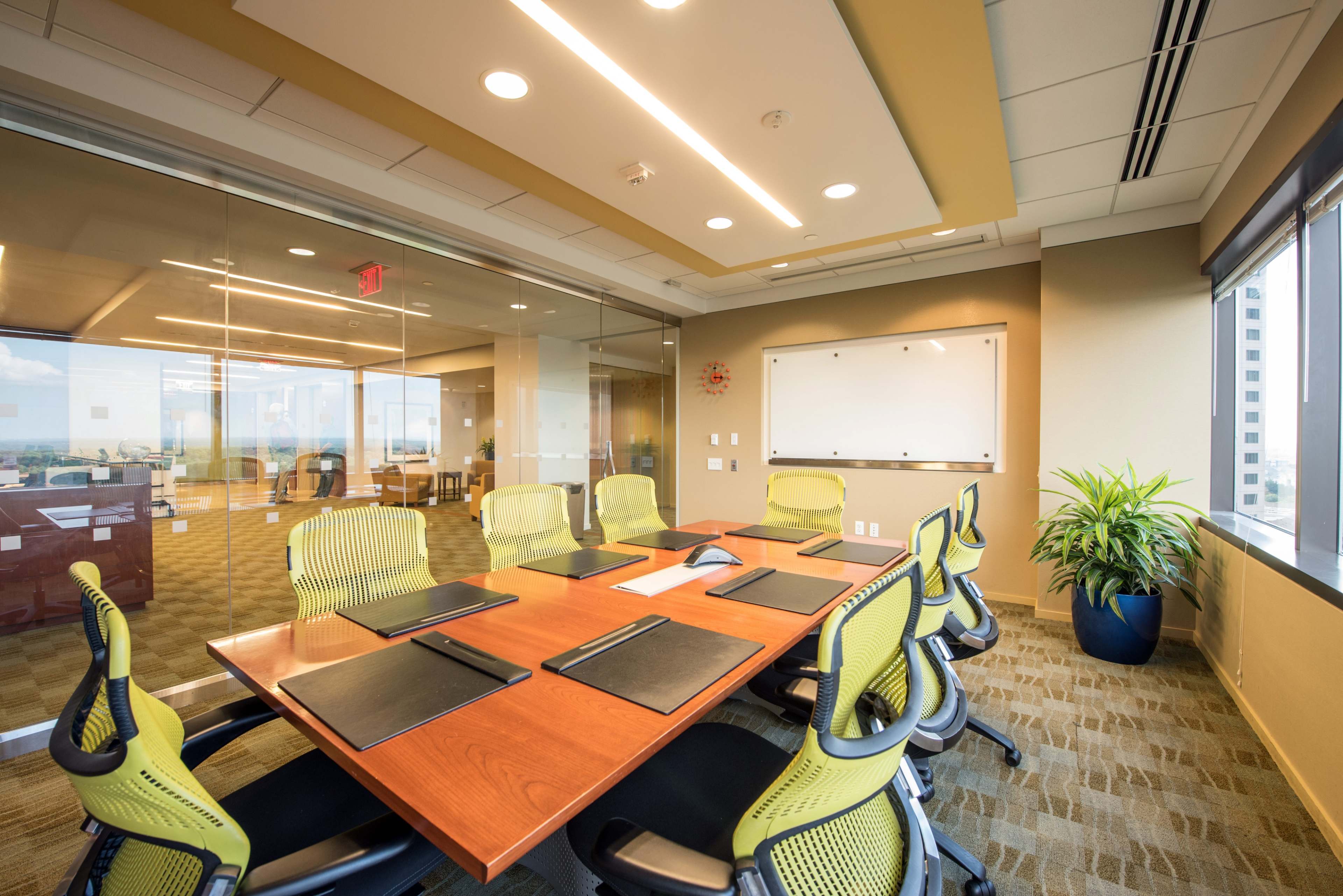 A glass-walled conference room features a wooden table surrounded by ergonomic chairs and equipped with writing materials.