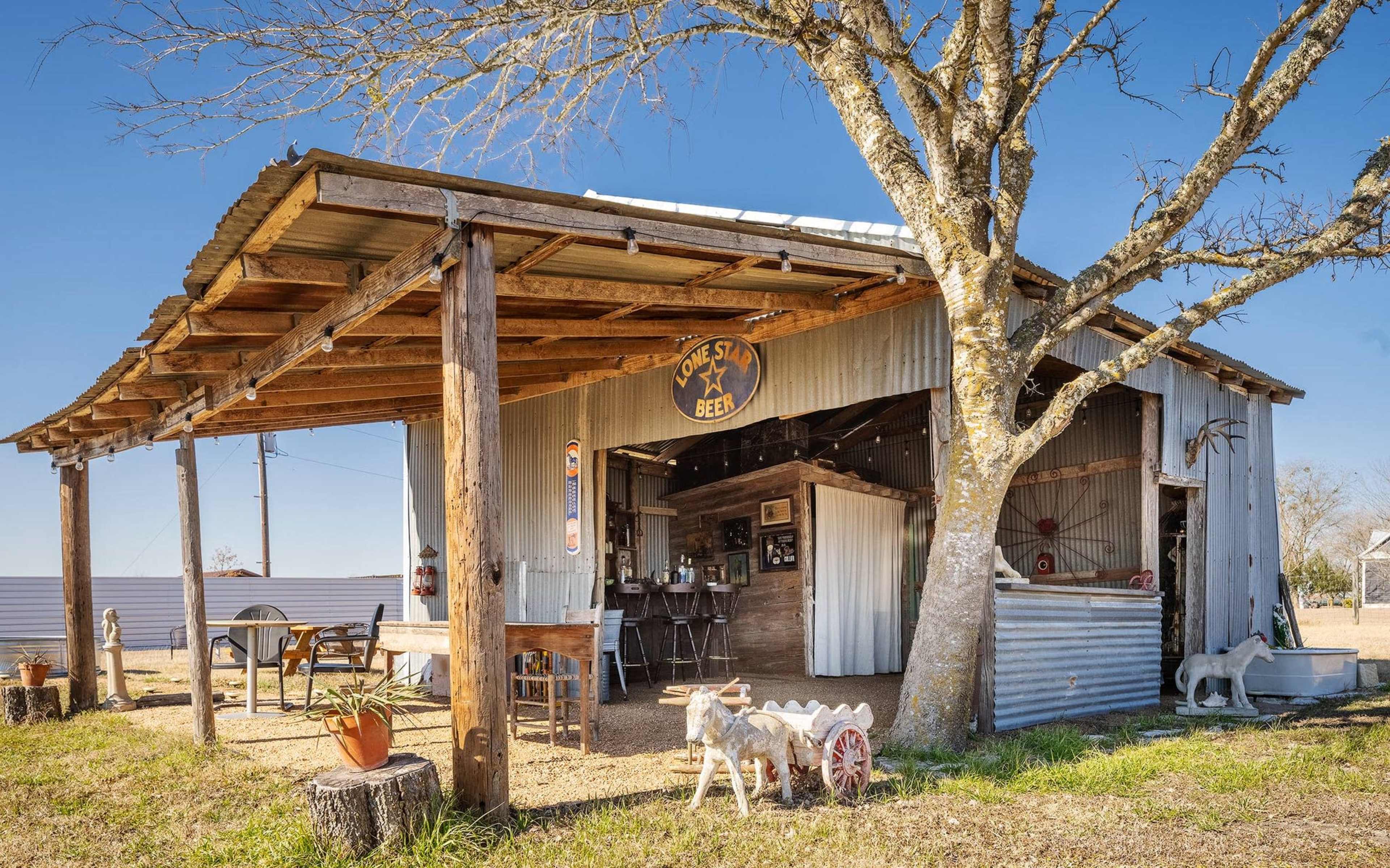 A rustic wooden shed with a tin roof and a porch features a sign that reads "Lone Star Beer" and includes outdoor seating with potted plants and a decorative cart.