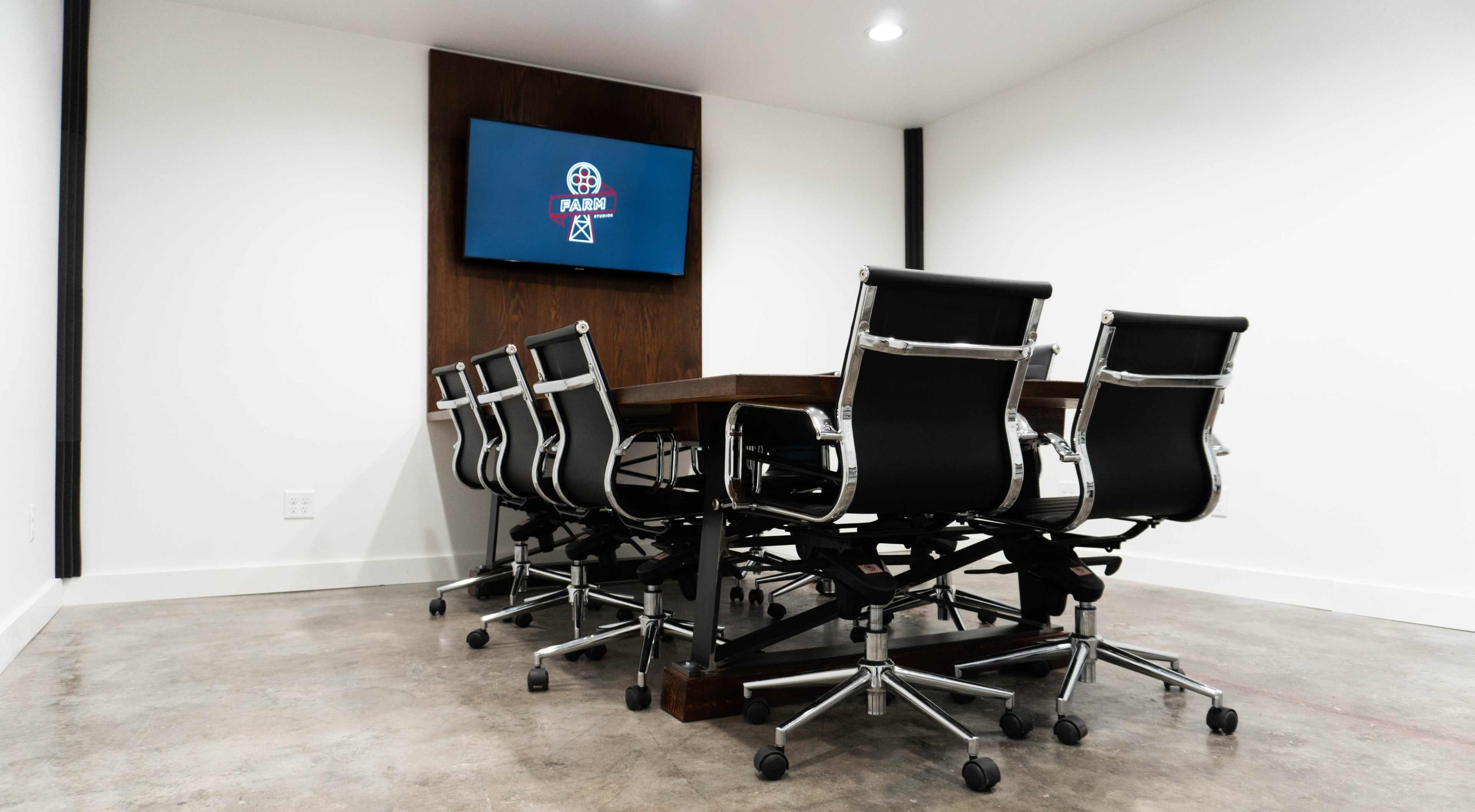 A conference room features a large wooden table surrounded by eight black rolling chairs, with a television mounted on the wall displaying a logo.