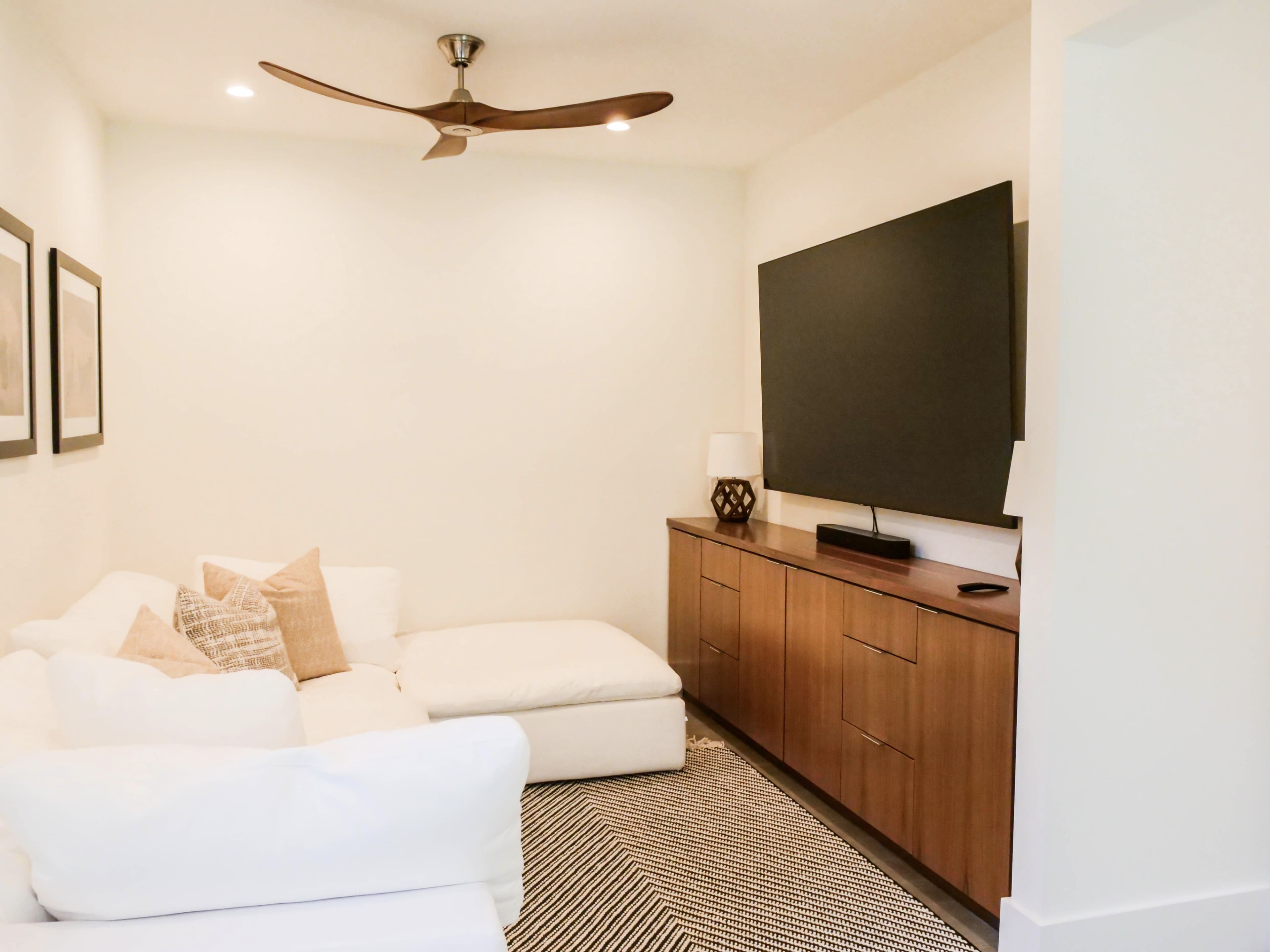 A living room with a white sectional couch, a wooden TV cabinet, and a large flat-screen television mounted on the wall.