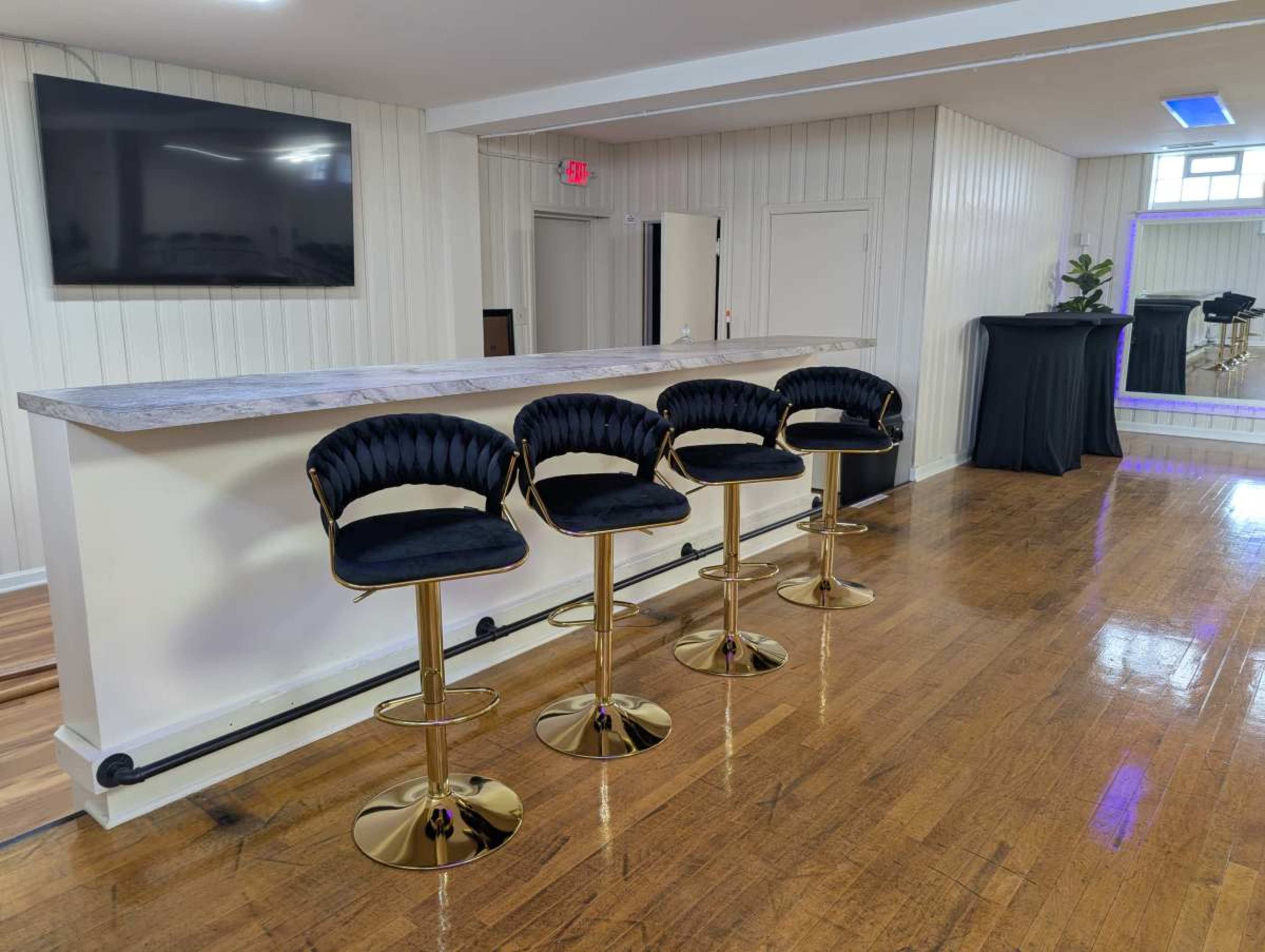 The image shows a modern bar area with four black and gold bar stools lined up in front of a marble counter and a wall-mounted television.