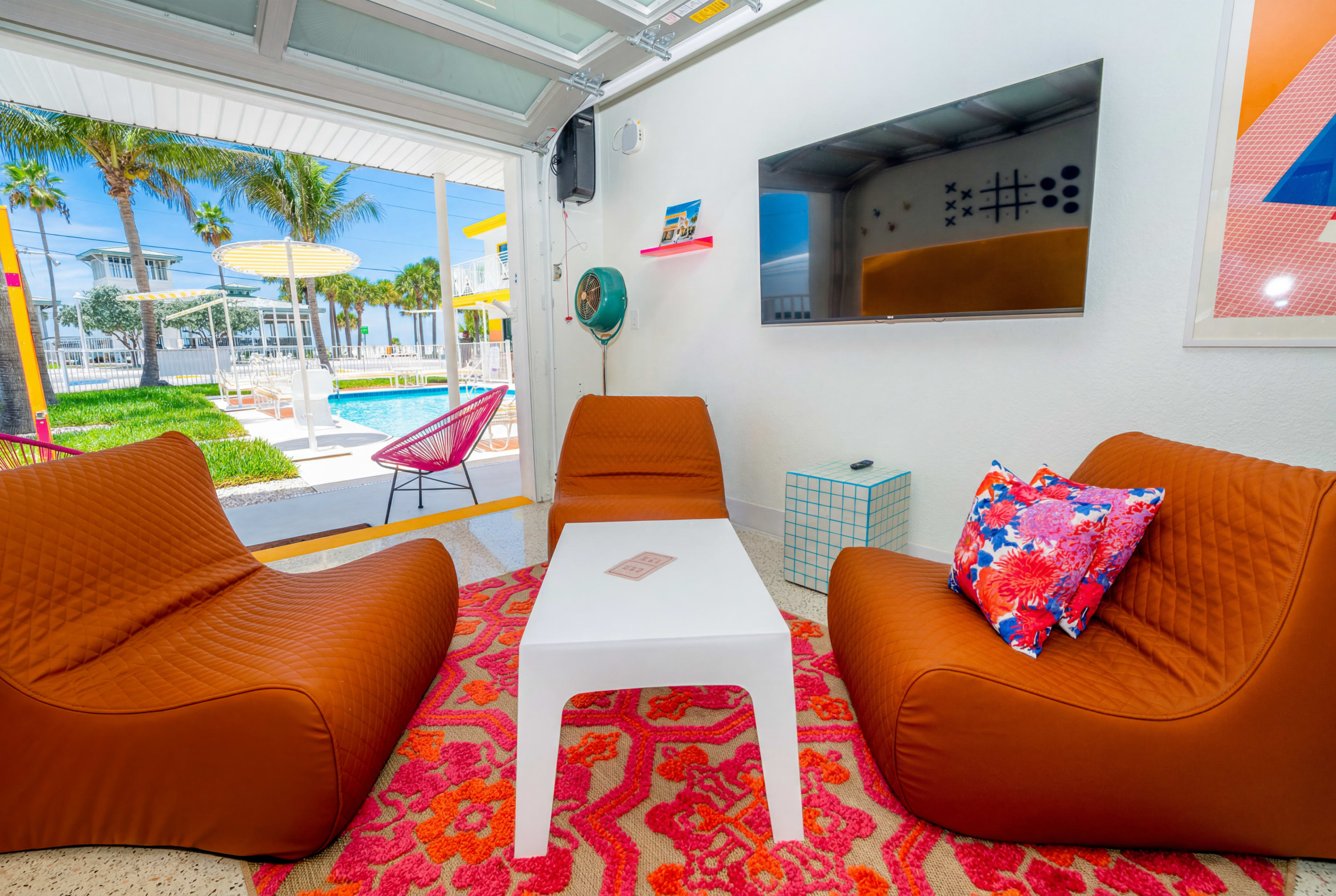 A modern lounge area features two brown beanbag chairs, a white table, and a colorful area rug, with a view of a pool and palm trees outside.