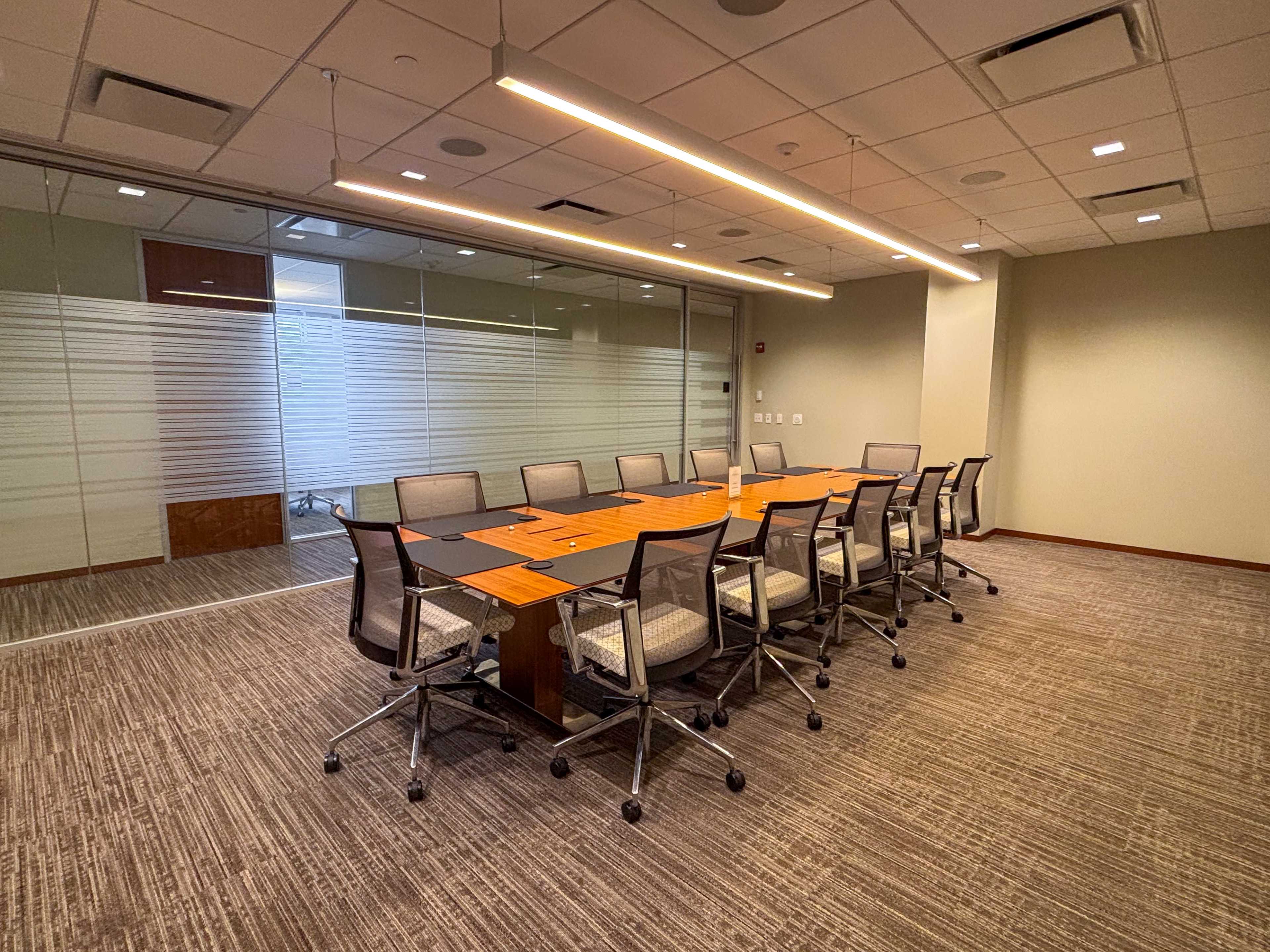 The image shows a modern conference room with a long table surrounded by ergonomic chairs and glass-paneled walls.