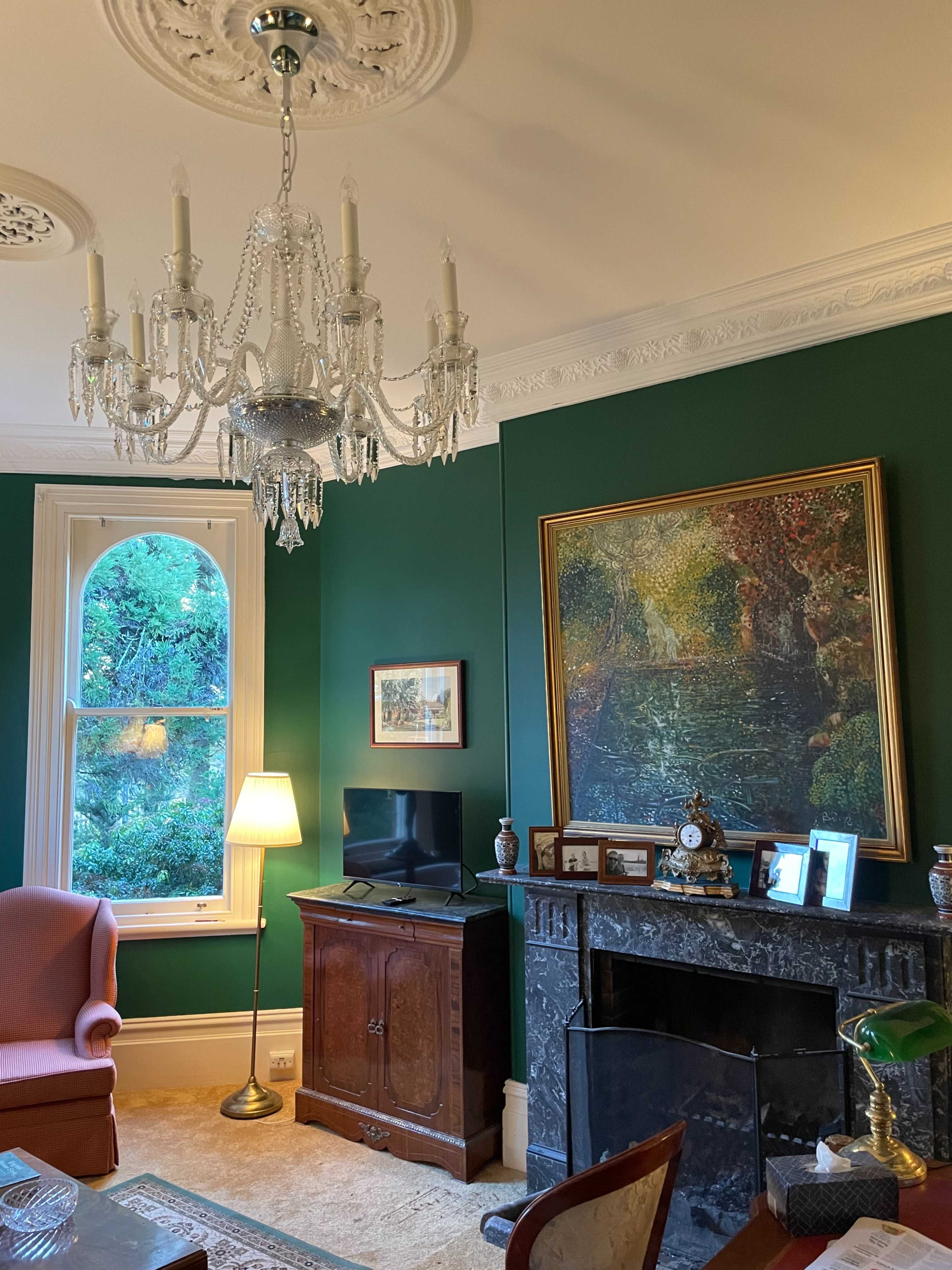 A room with green walls, a chandelier, a fireplace, a television, a decorative painting, and a lamp beside a pink armchair.