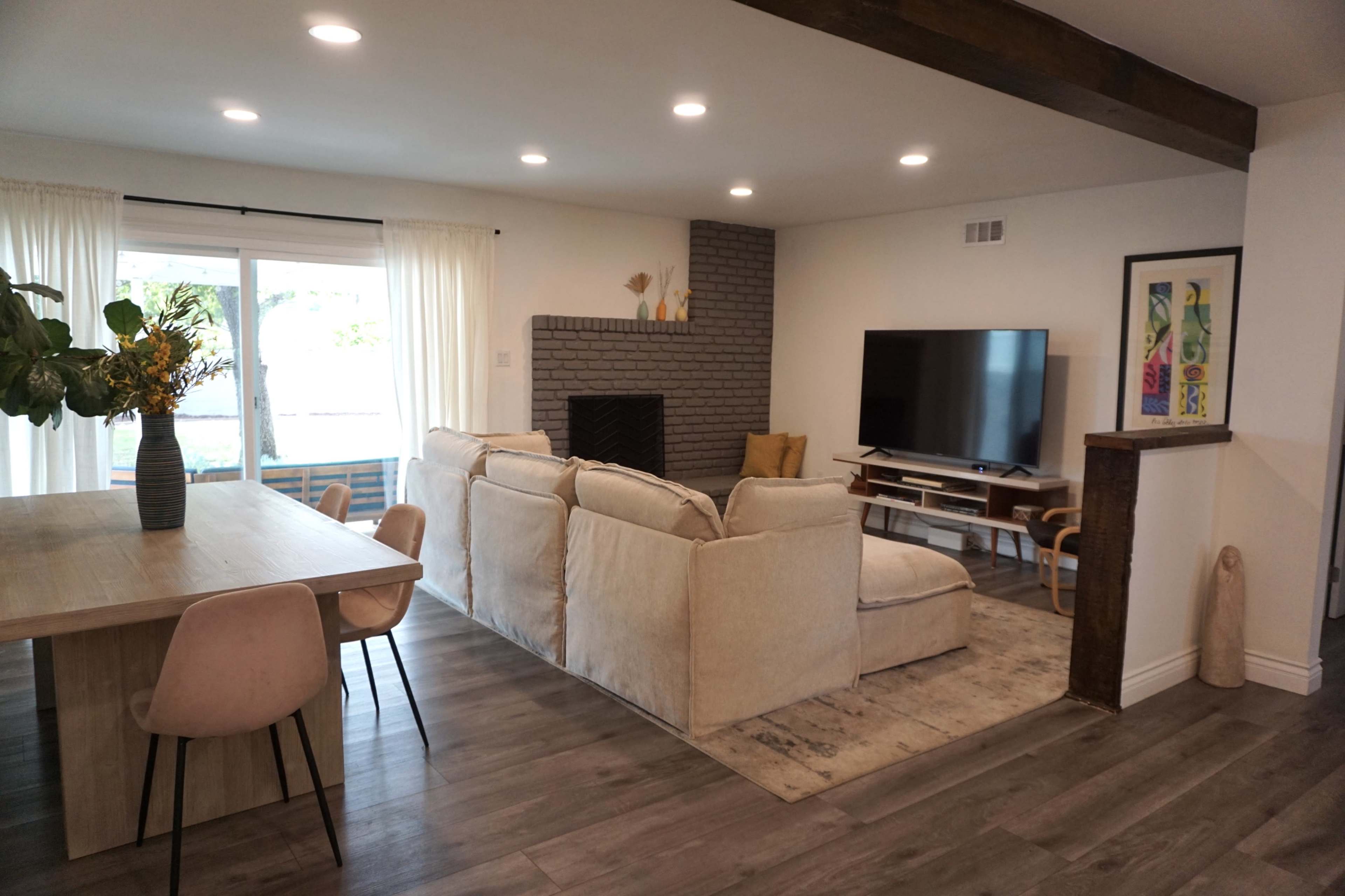 The image shows a modern living room with a beige sectional sofa, a television mounted on the wall, and a dining table with four chairs.
