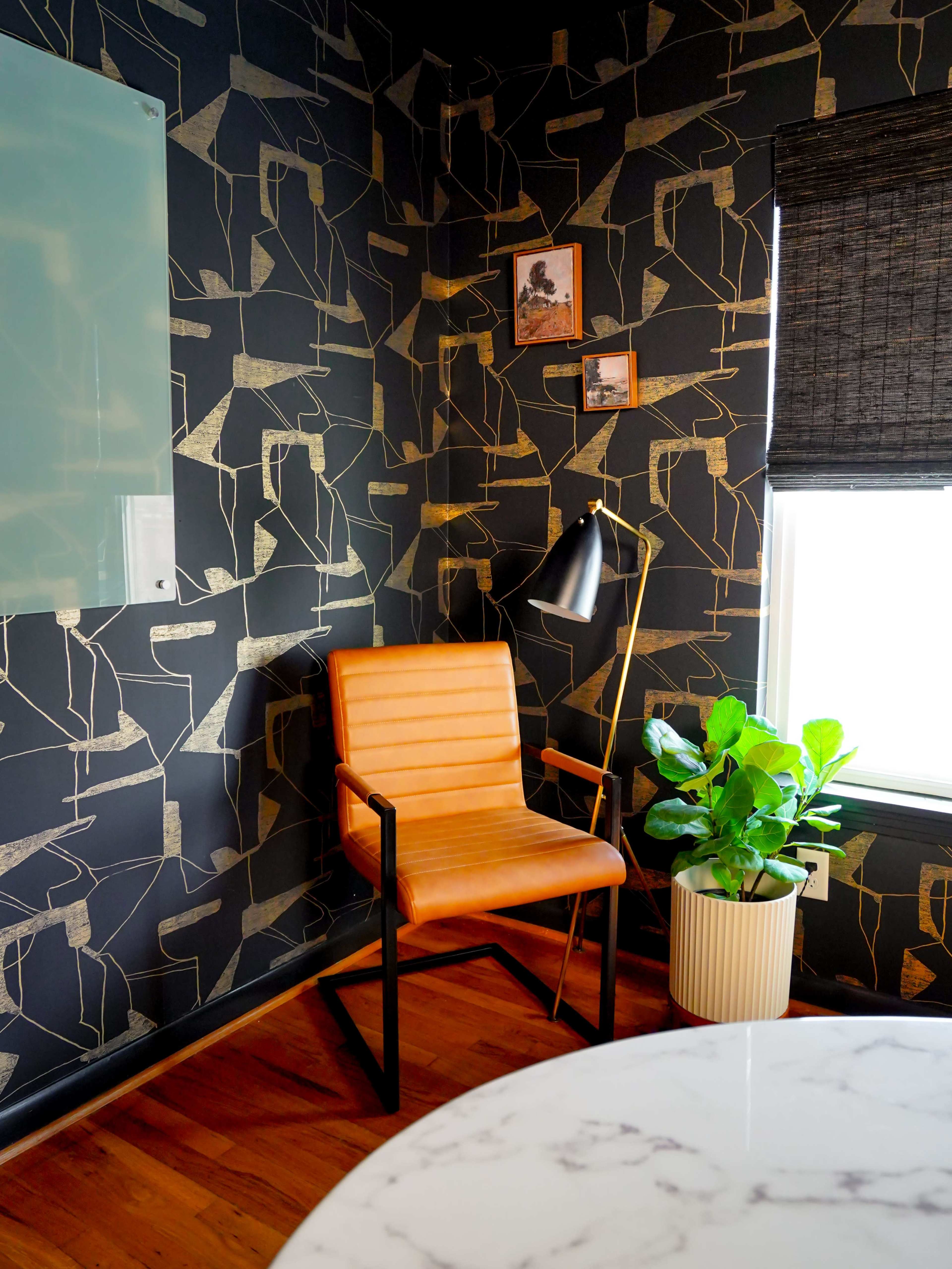 A modern corner office features a tan leather chair next to a floor lamp, with a large plant and framed pictures on the textured black wallpapered wall.