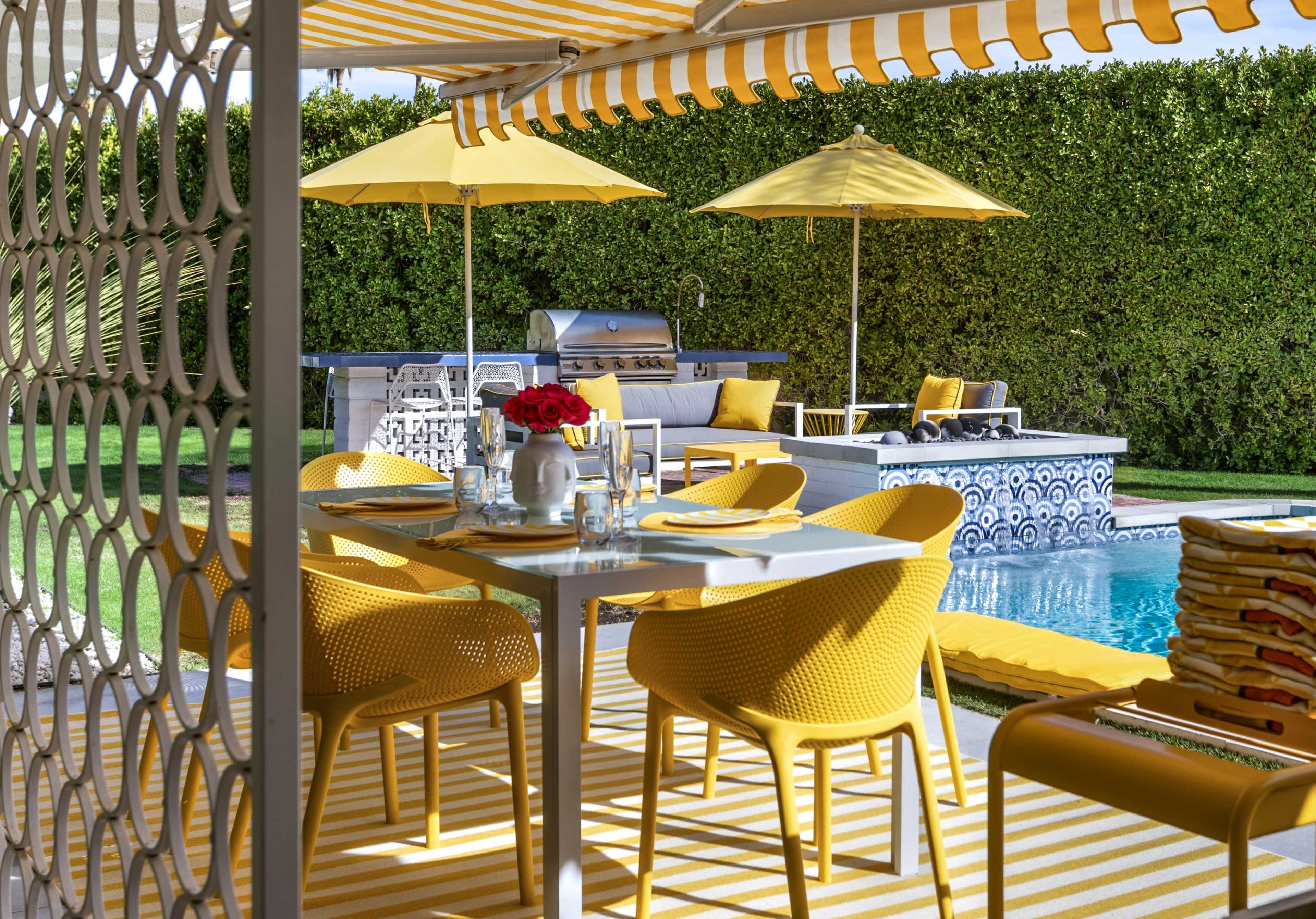 A bright outdoor patio features yellow furniture, a dining table, and a swimming pool surrounded by a lush green hedge.