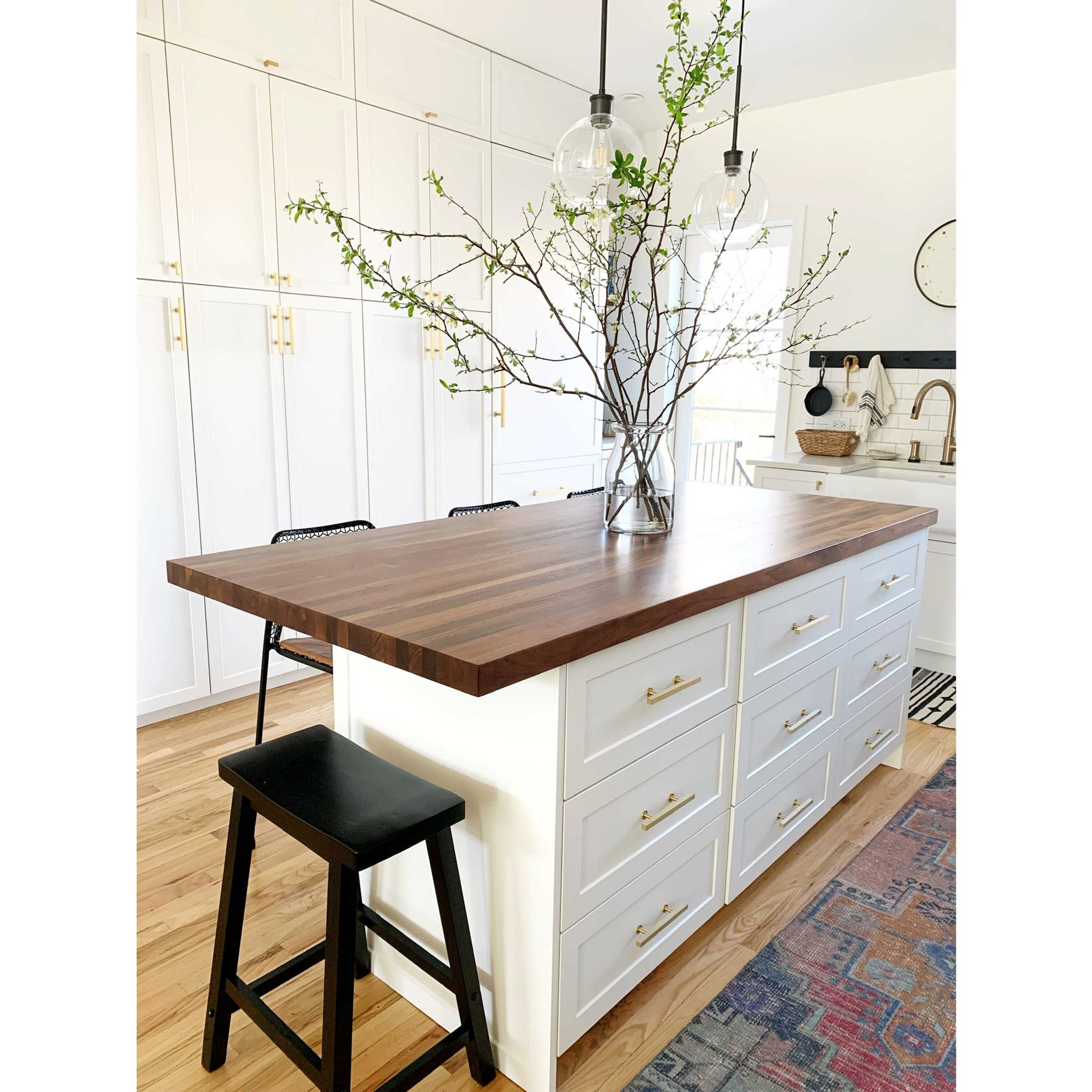 A modern kitchen features a central island with a dark wooden countertop, white drawers, and a vase containing branches, complemented by pendant lighting above.