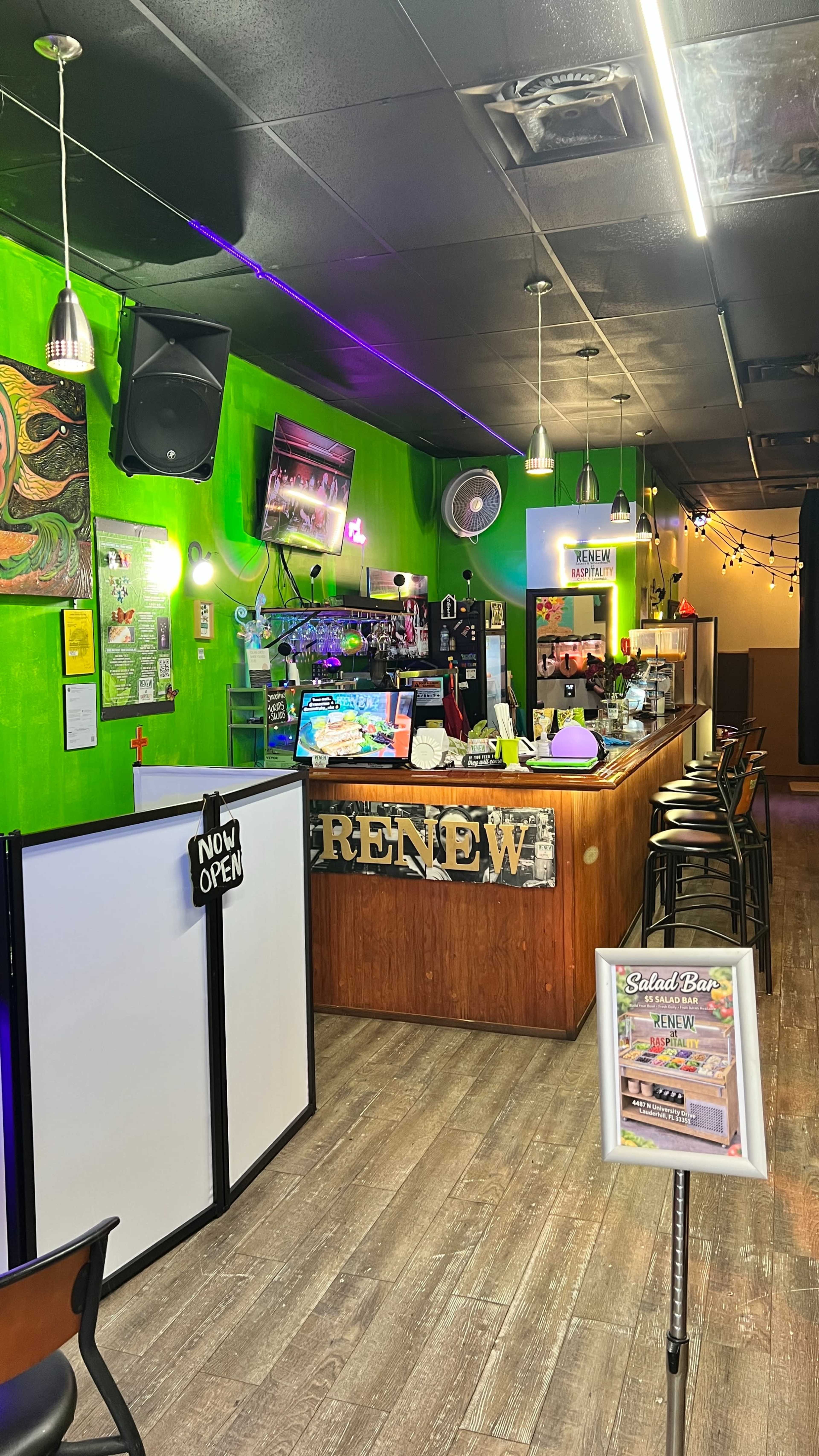 The image shows a brightly colored café or bar interior with a wooden counter, seating arrangements, and a sign indicating that it is now open.