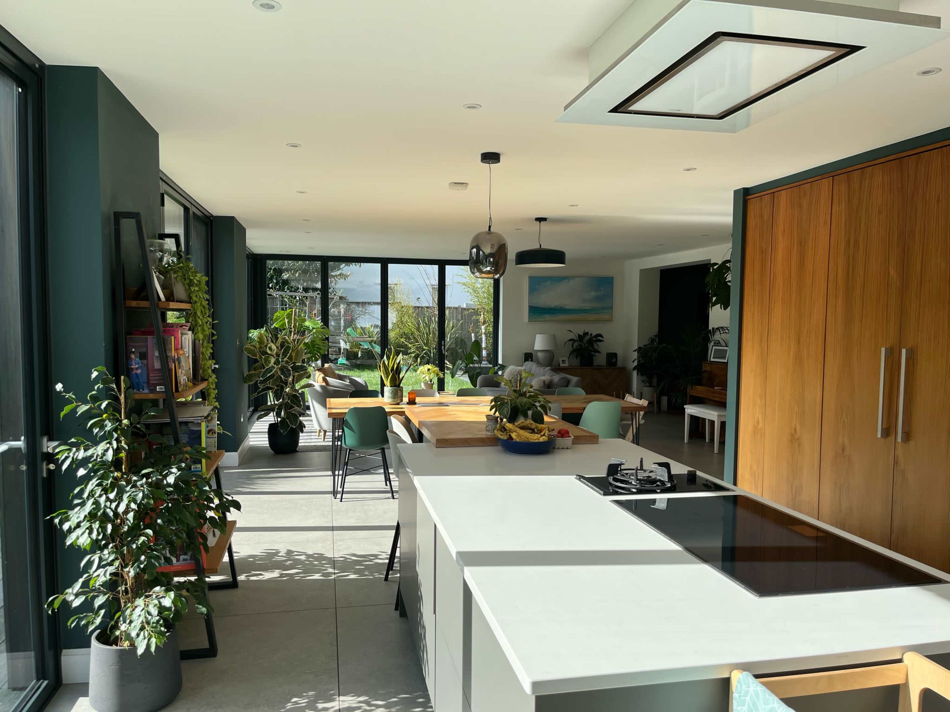 A modern kitchen with a central island features an open layout connecting to a dining area filled with plants and natural light.