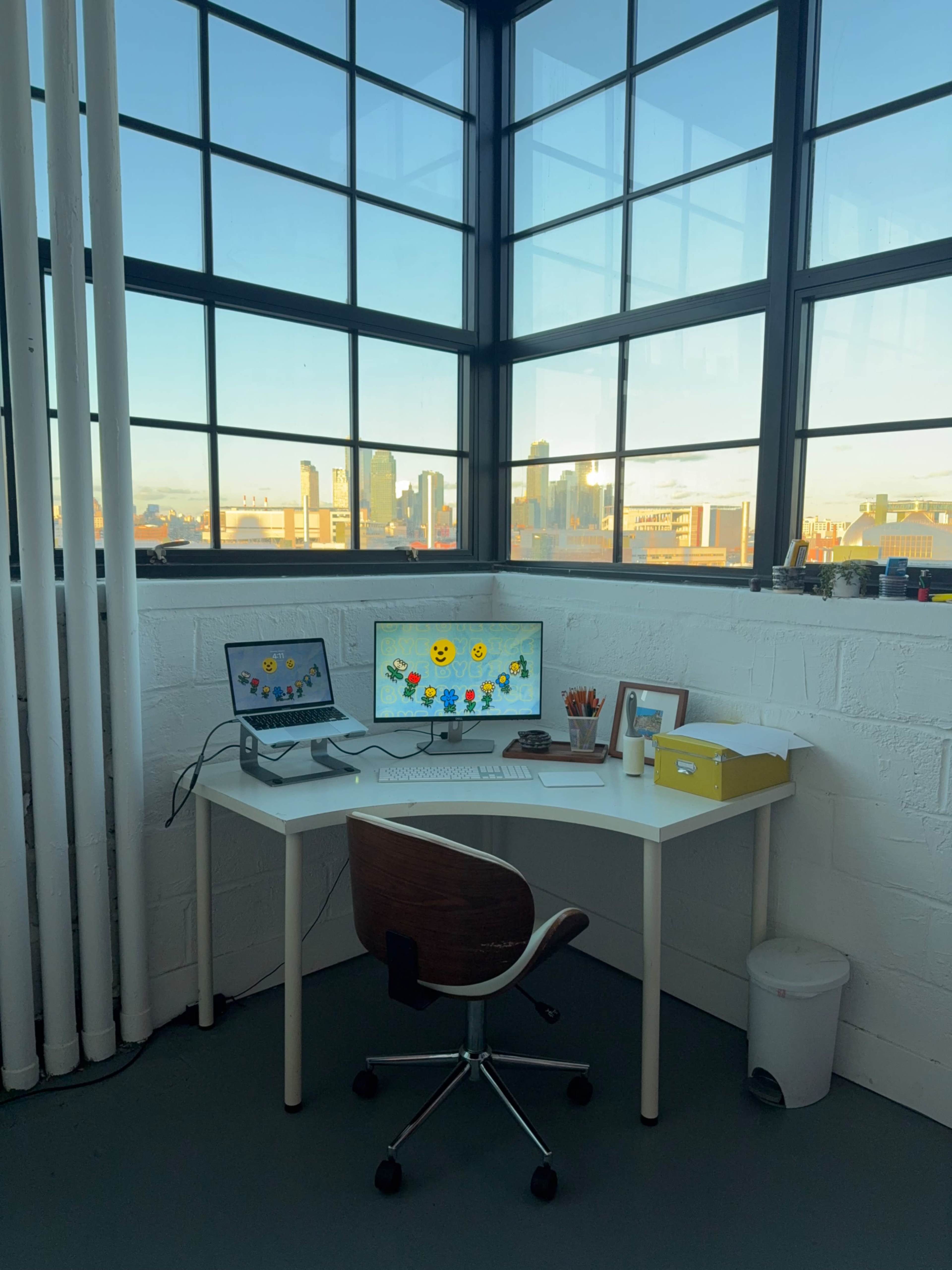 A modern workspace features a white desk with two monitors, a chair, and a wastebasket, illuminated by natural light streaming through large windows with a city skyline visible in the background.