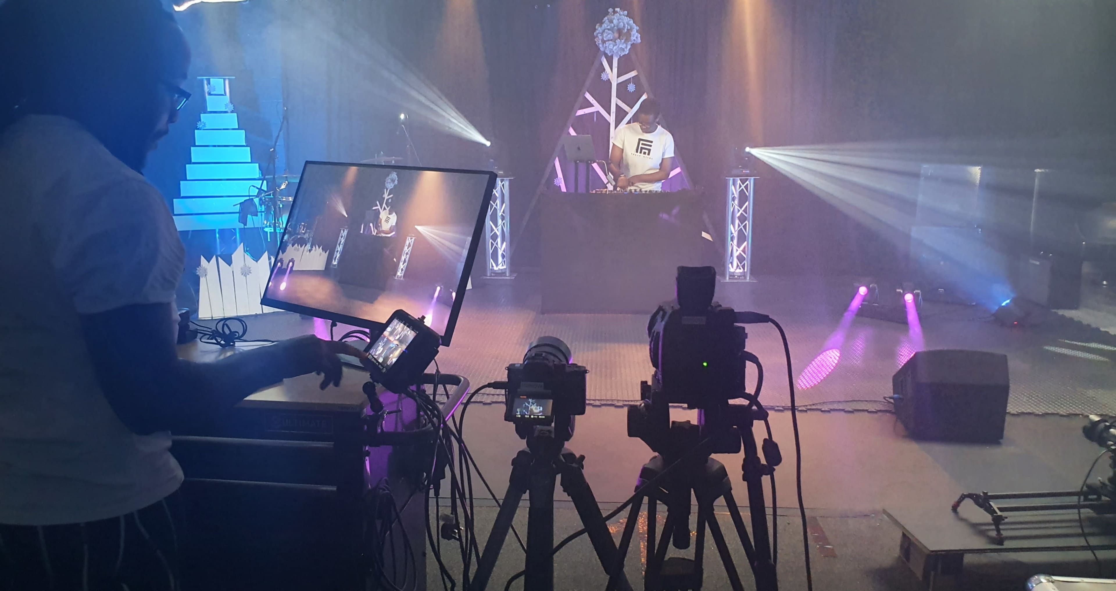 A DJ performs on stage with multiple cameras and lighting effects in a dimly lit venue.