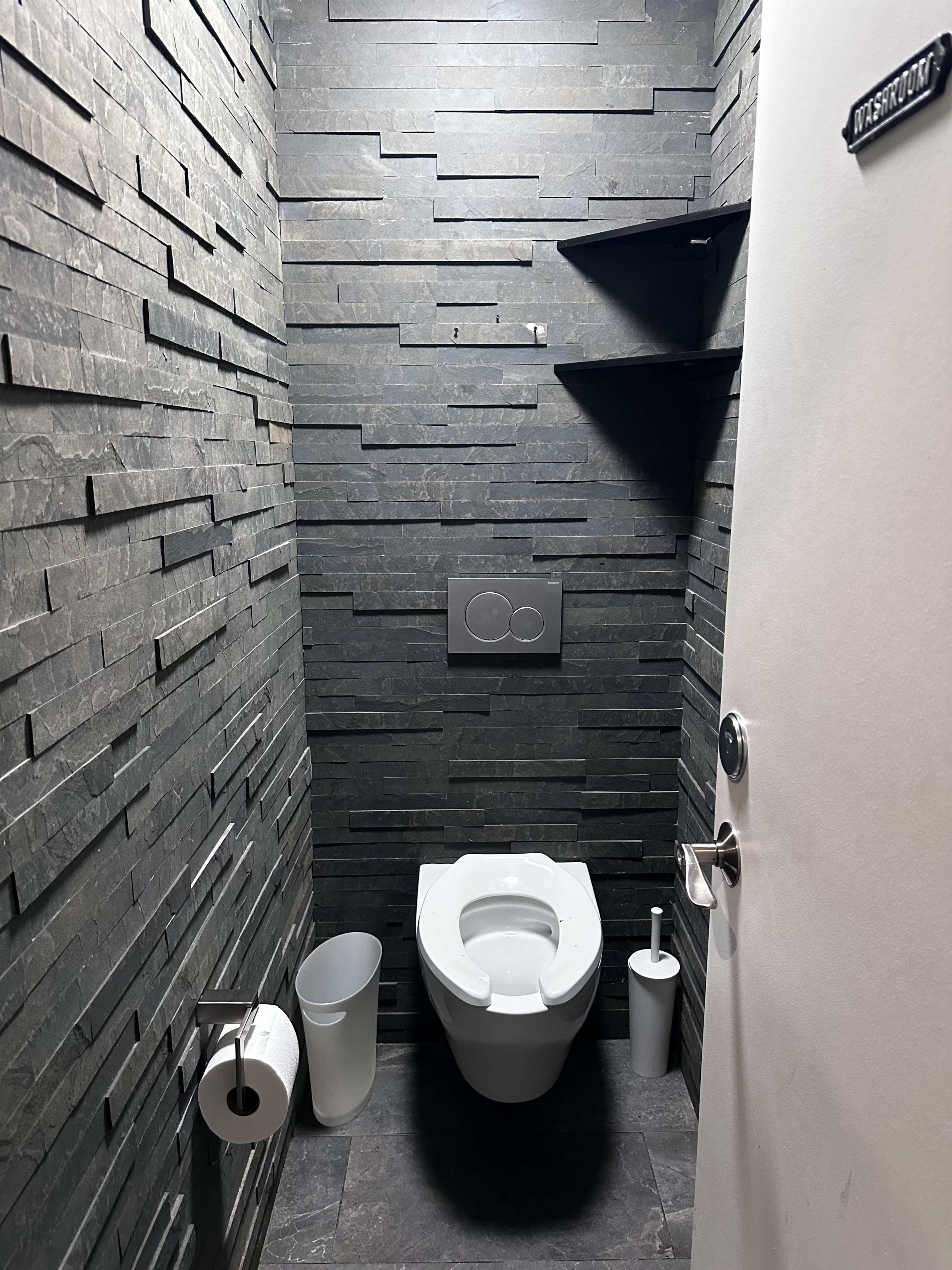 The image shows a small restroom with a dark stone wall, a modern toilet, and minimal shelf storage.