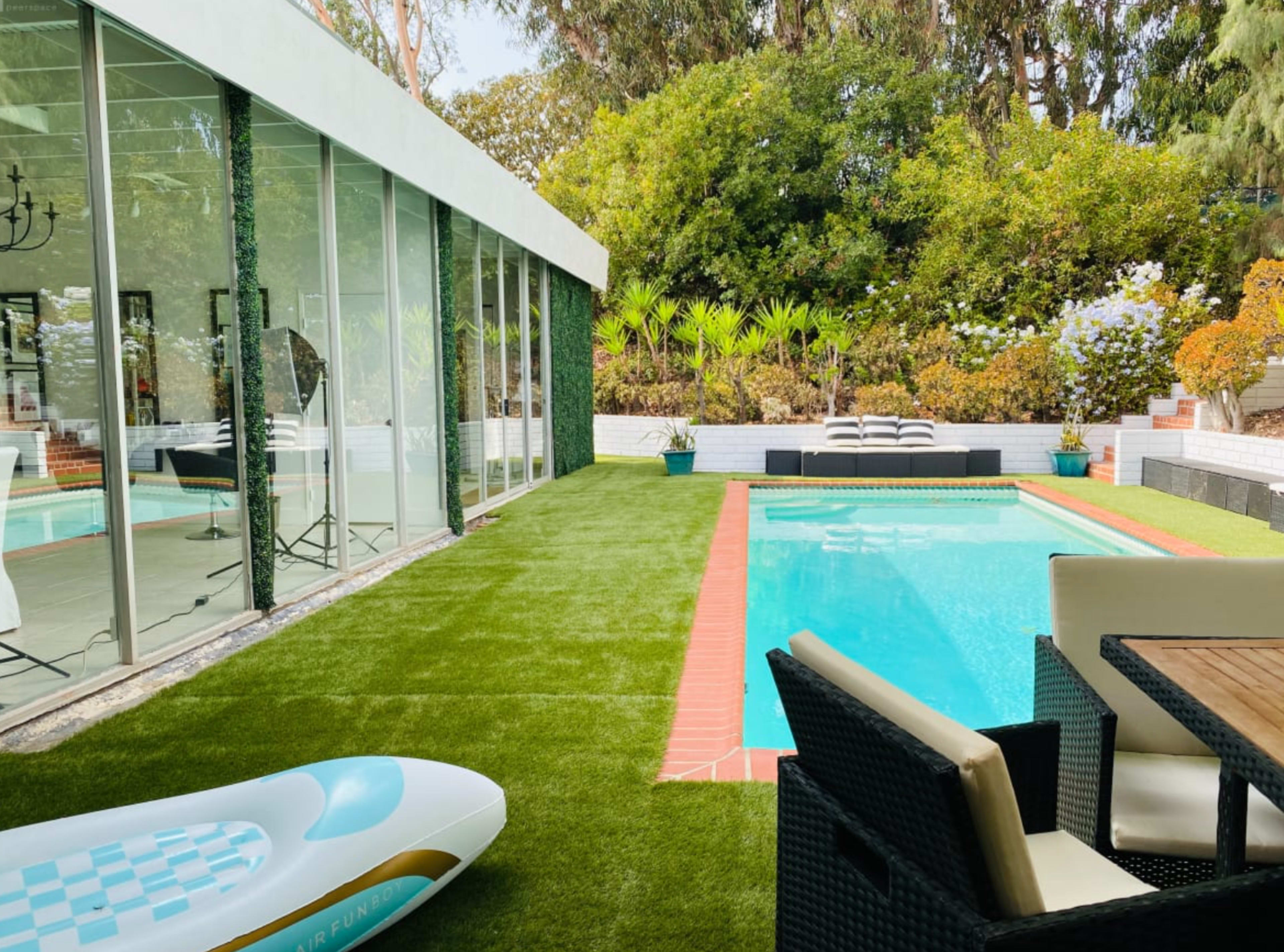 A modern outdoor space featuring a swimming pool surrounded by artificial grass, lounge chairs, and a glass-walled house.
