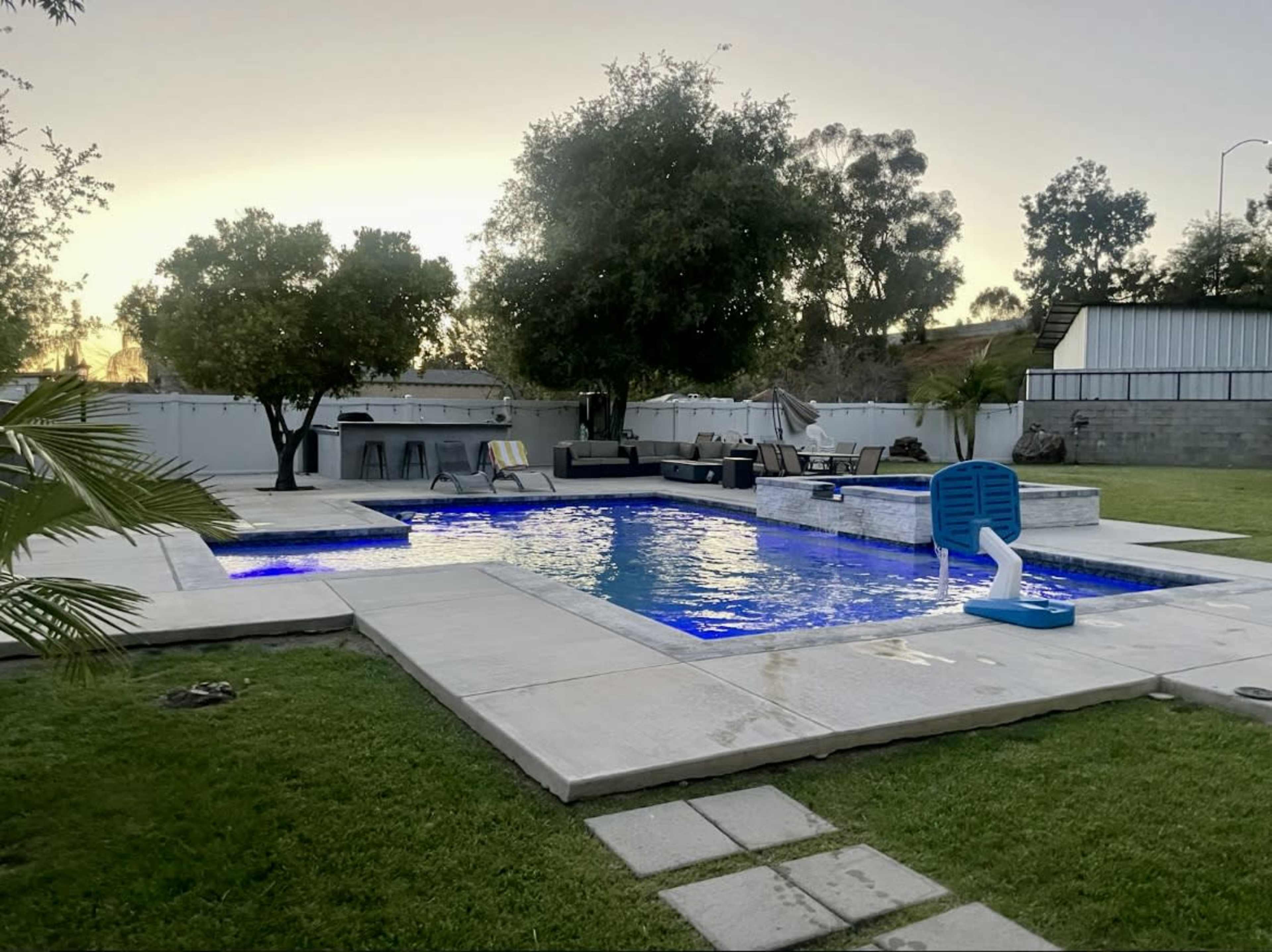 A rectangular swimming pool with blue lighting and a shallow area is surrounded by a patio, lounge chairs, and grass, set against a backdrop of trees and a house.
