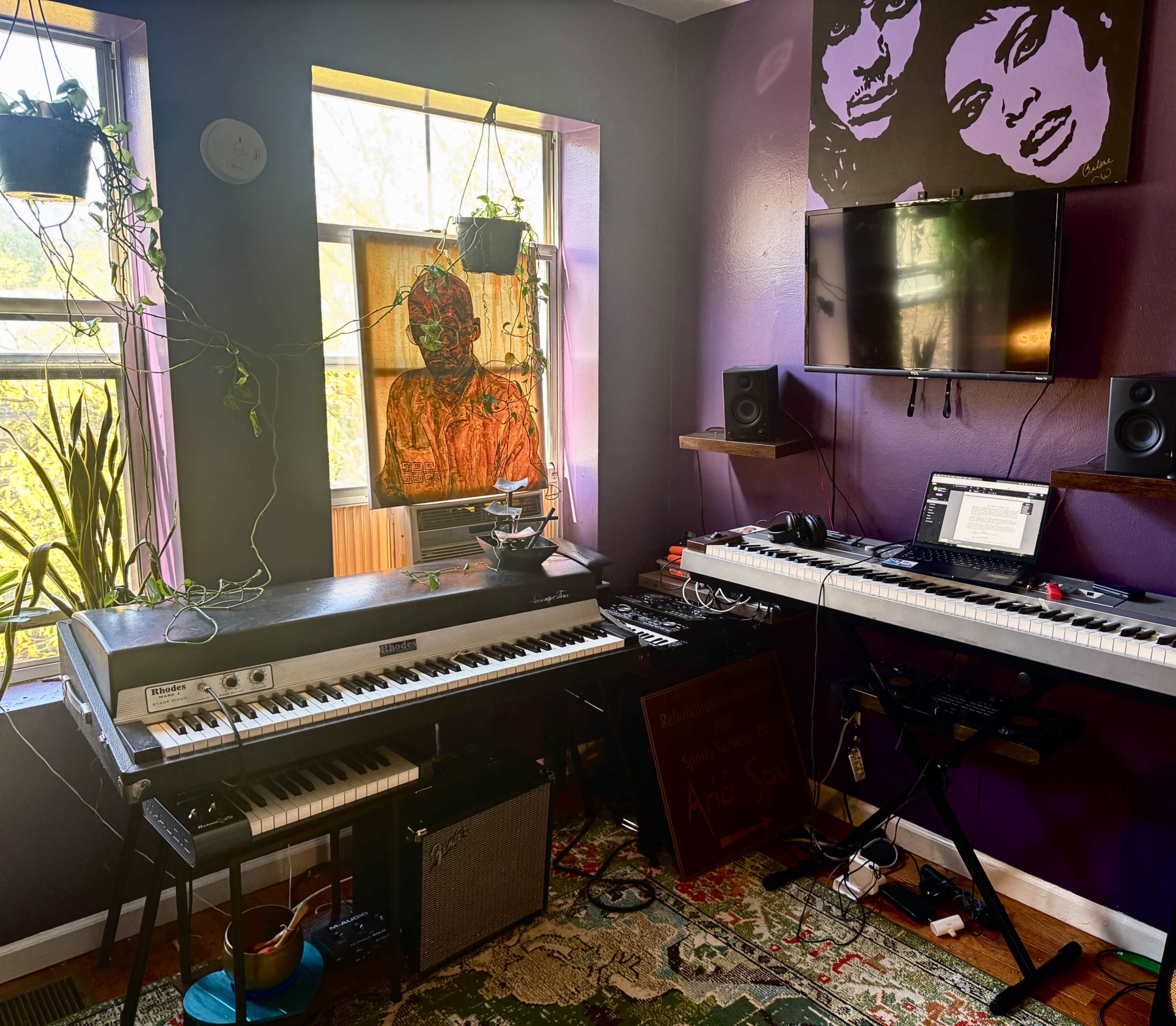 The image shows a vibrant music studio featuring electronic keyboards, a computer setup, framed artwork on the walls, and plants in the windows.