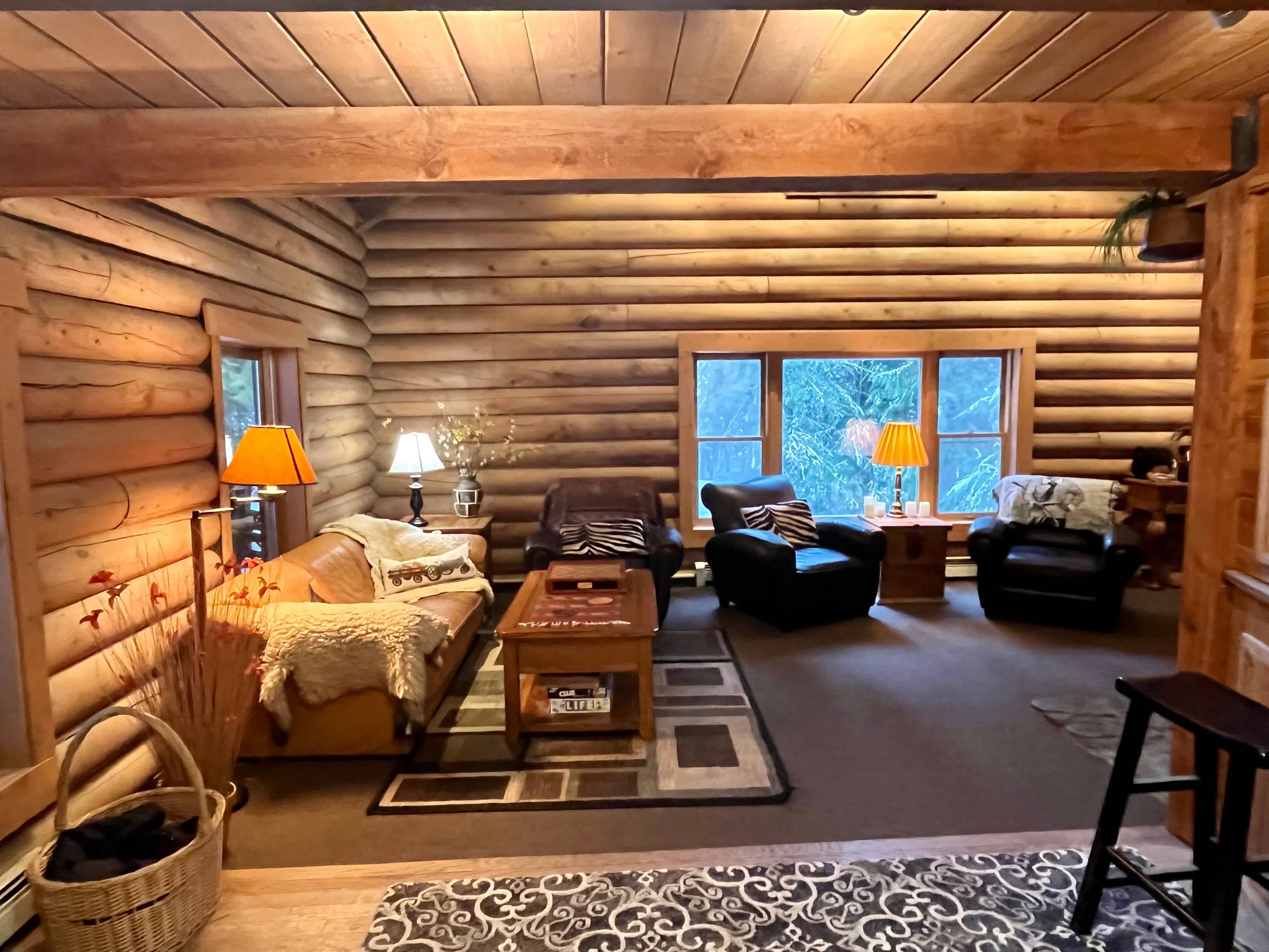 A cozy log cabin living room featuring a mix of seating arrangements, wooden accents, and large windows offering a view of the outdoors.