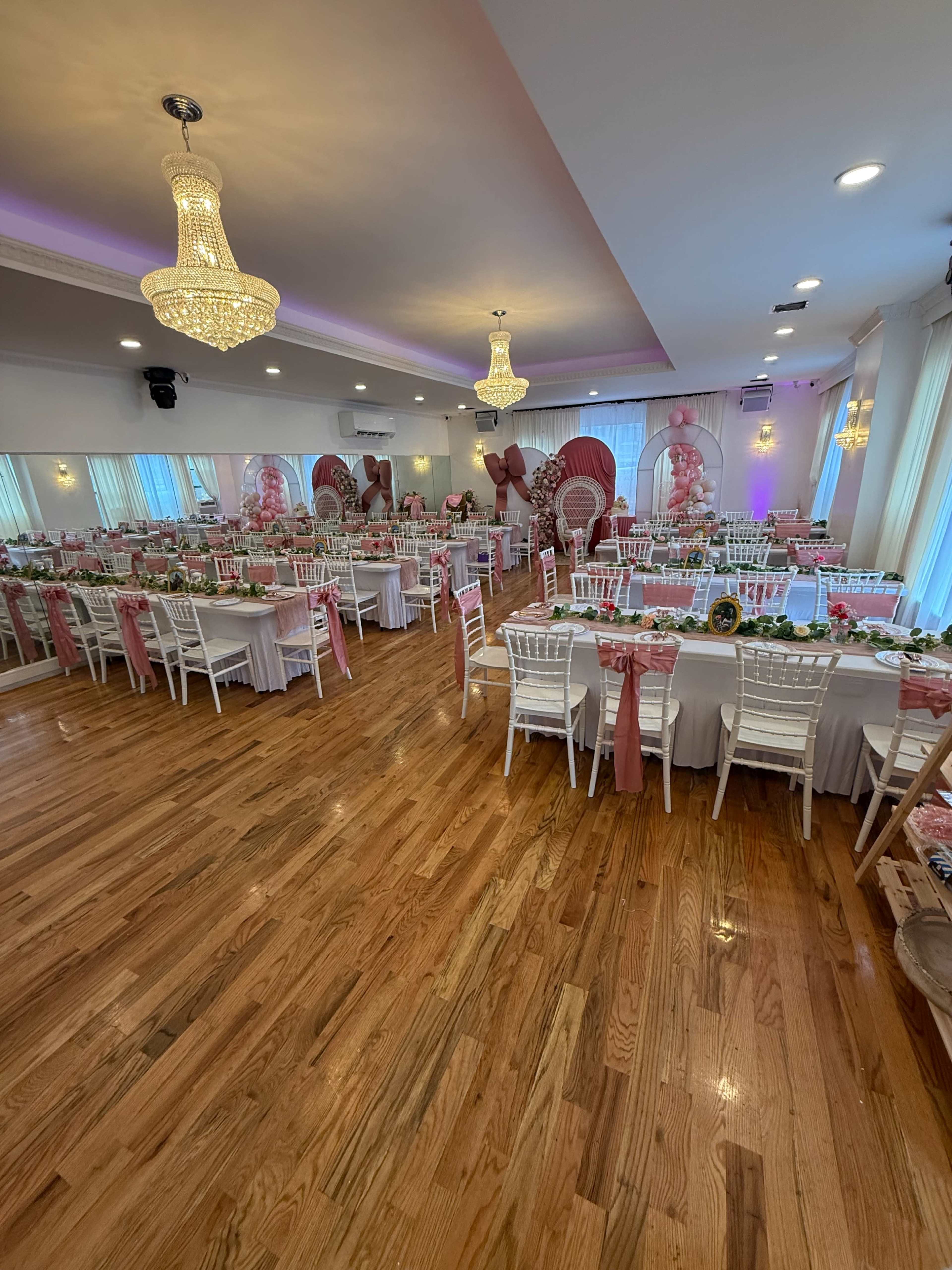 The image shows a decorated banquet hall with white tables set for an event, featuring pink accents, floral centerpieces, and elegant chandeliers.