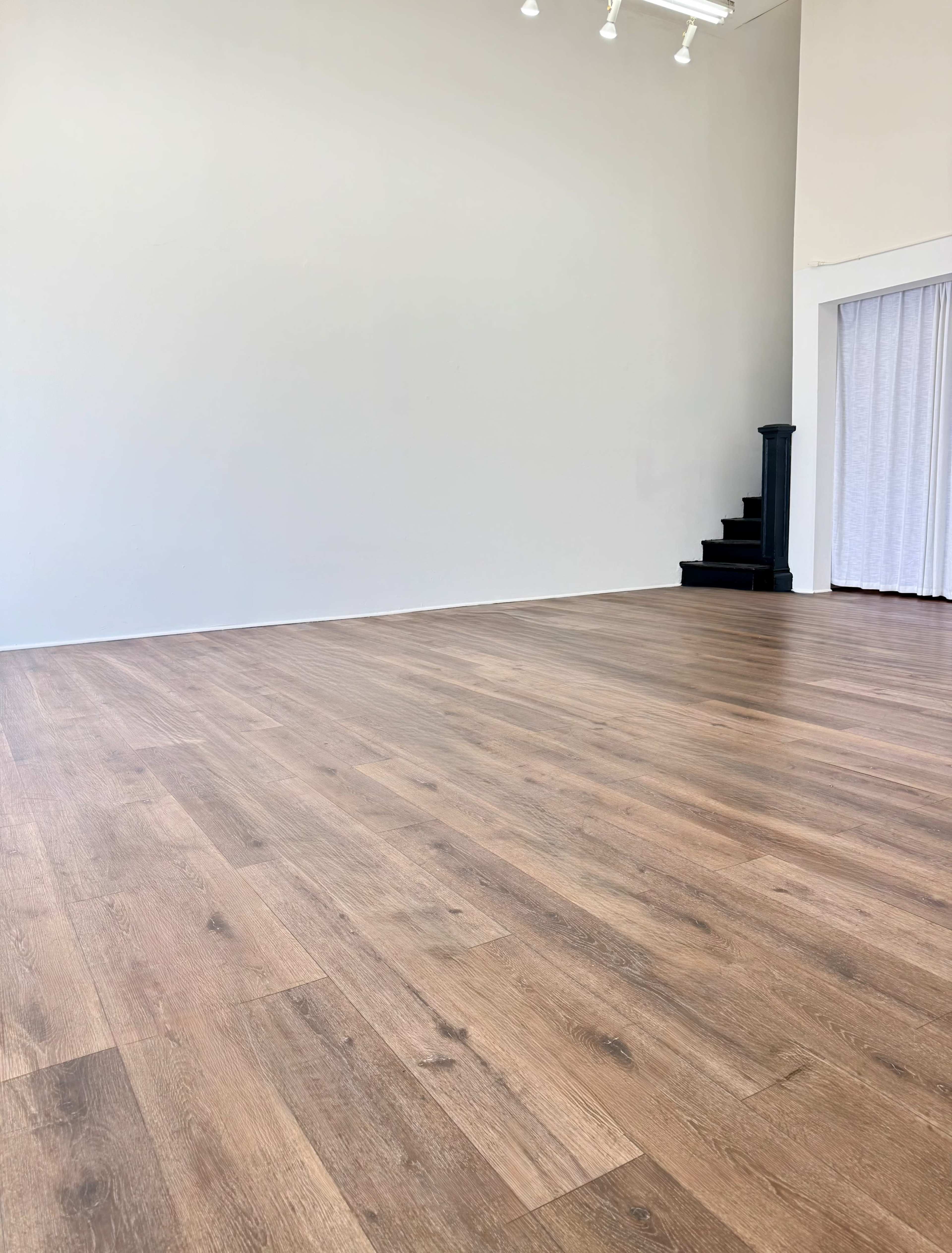 The image shows a spacious room with wooden flooring, featuring a plain white wall and a staircase in the background.